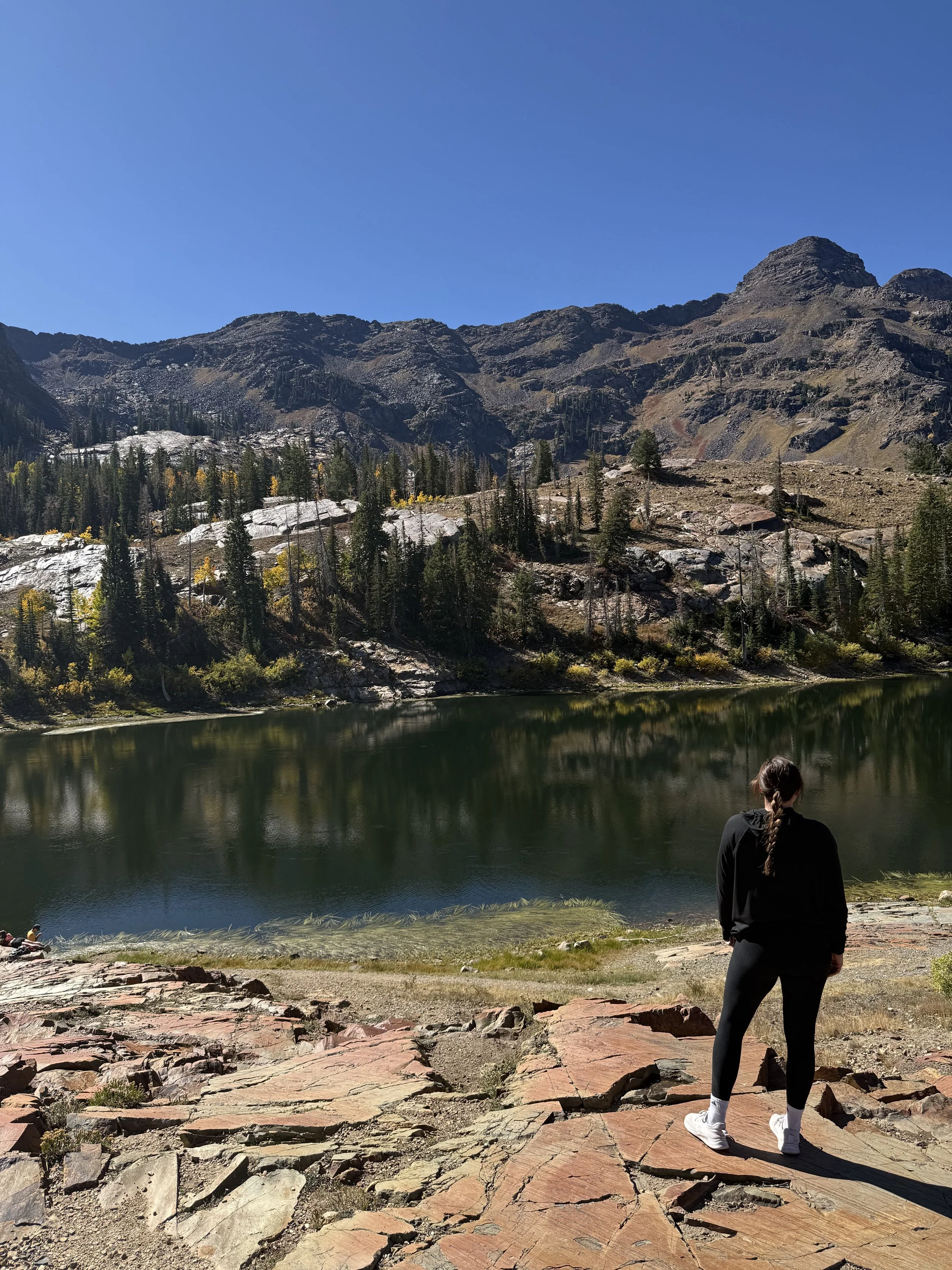 A person standing on rocky ground near a calm lake, surrounded by forested mountains under a clear blue sky.