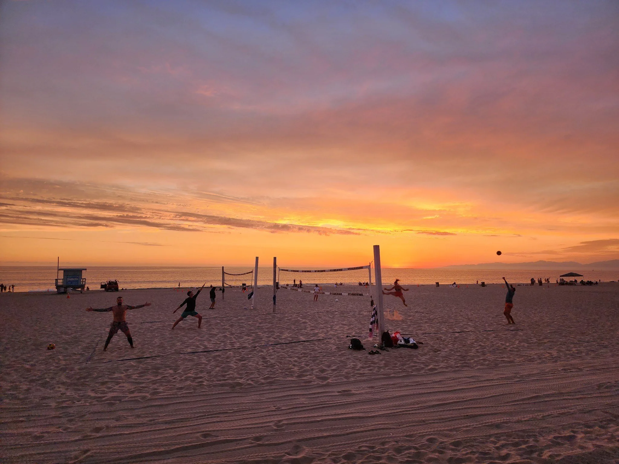 People playing beach volleyball on the sand during sunset at the beach with the ocean in the background.
