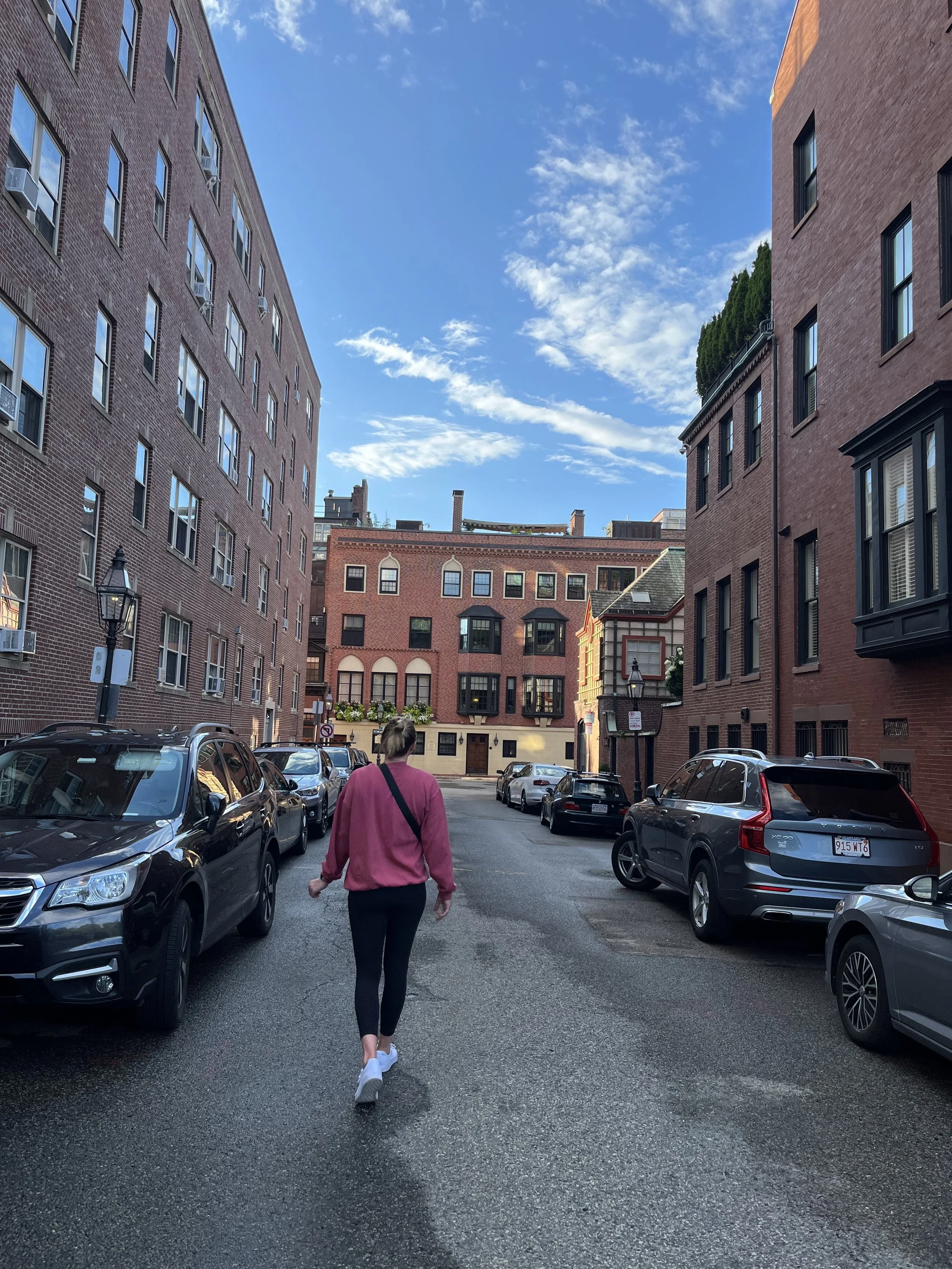 A woman walking down a city street with parked cars on both sides and tall brick buildings on either side under a partly cloudy sky.