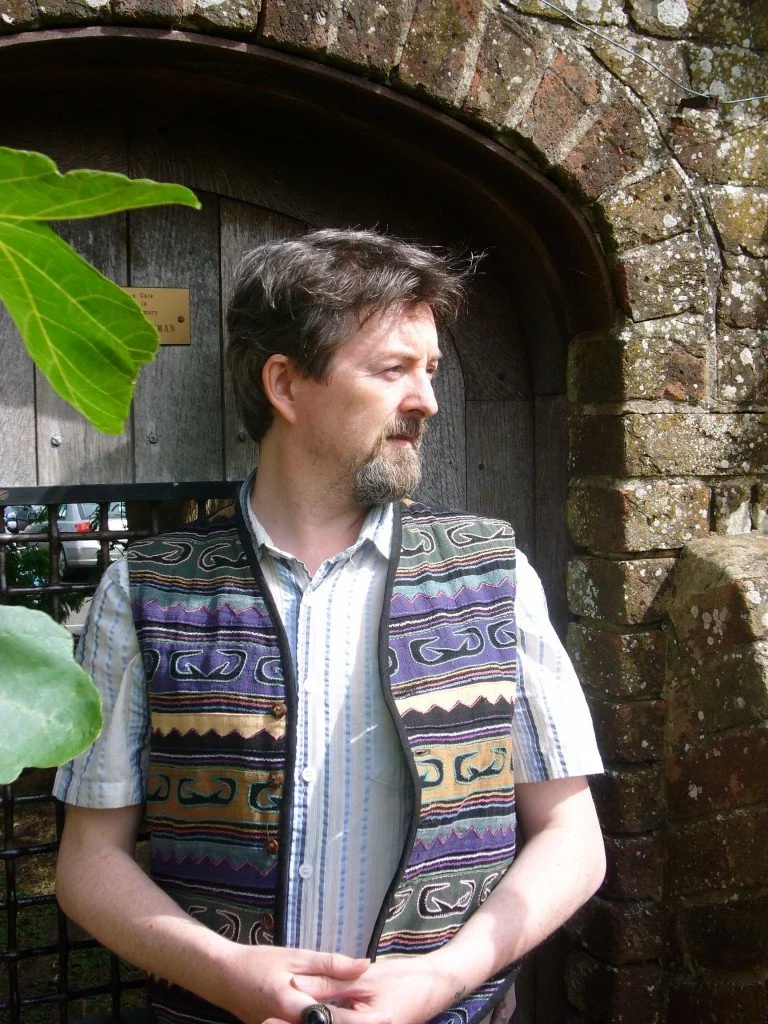 Matthew Levi Stevens con barba y cabello rizado, usando un chaleco con patrones coloridos, de pie junto a una puerta de madera en un entorno exterior con ladrillos y vegetación.
