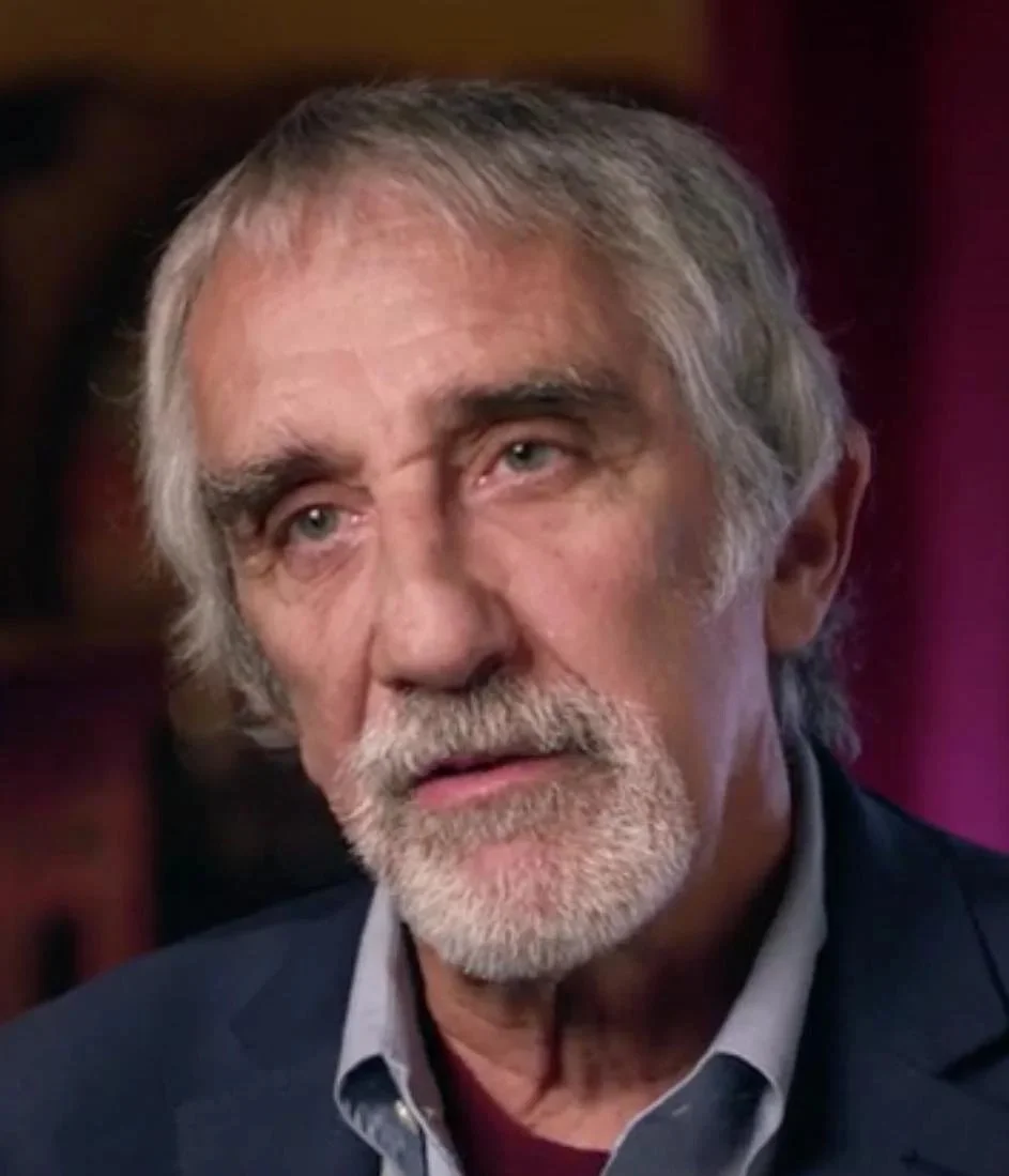 James Wasserman con cabello y barba gris, expresión pensativa, vestido con traje y camisa, fondo borroso.