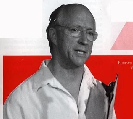 Retrato de Ramsey Dukes en blanco y negro de un hombre con gafas, camisa blanca y sonriendo, sostenido un libro en una mano, con fondo con áreas en rojo y gris.