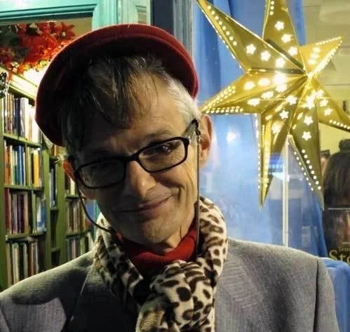 Jake Stratton-Kent con gafas y bufanda de leopardo en un ambiente con una estrella decorativa iluminada y estantes llenos de libros.