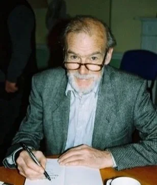 Mike Mitchell con barba y gafas escribiendo en un cuaderno, lleva puesto un saco gris y camisa azul, en un ambiente interior.