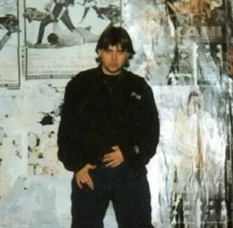 Spencer Kansa frente a un muro cubierto de posters y graffiti en un entorno urbano.