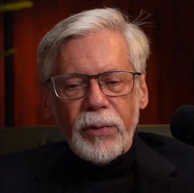 Peter Levenda con barba y gafas, hablando o en entrevista en un ambiente interior con fondo de madera.