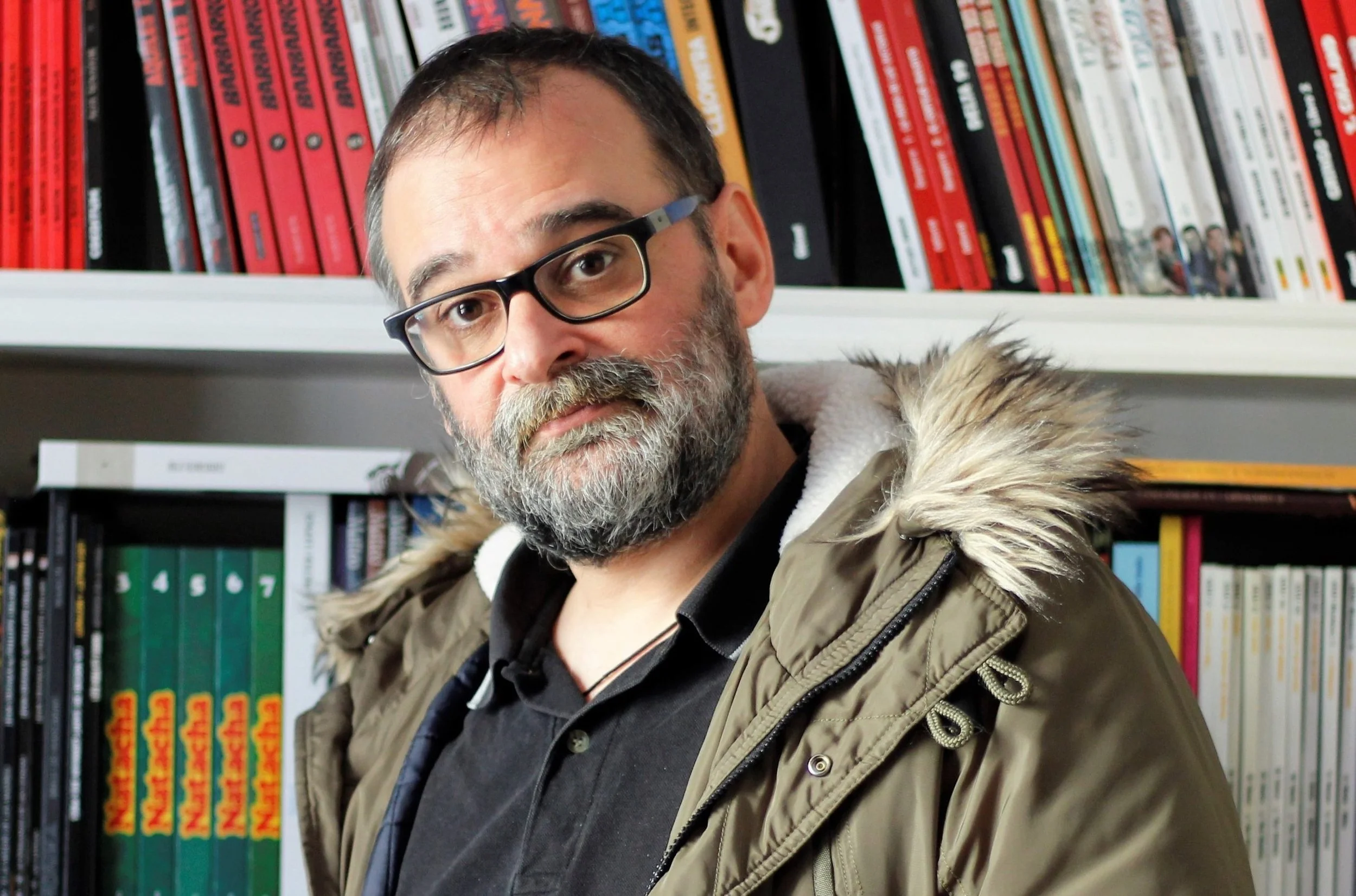 Alberto Ávila Salazar con barba, gafas y chaqueta con capucha de peluche, posando en una librería con estanterías llenas de libros.