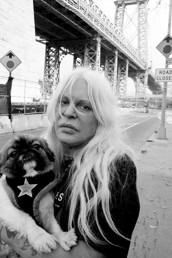 Genesis P-Orridge con cabello rubio largo sostiene un cachorro de raza bulldog en un entorno urbano, con un puente de fondo y señales de cierre de carretera en la calle.
