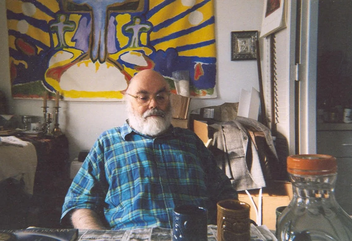 Michael Bertiaux con barba y gafas en una habitación, sentado frente a una mesa con tazas y botellas, fondo con arte abstracto colorido y objetos diversos.