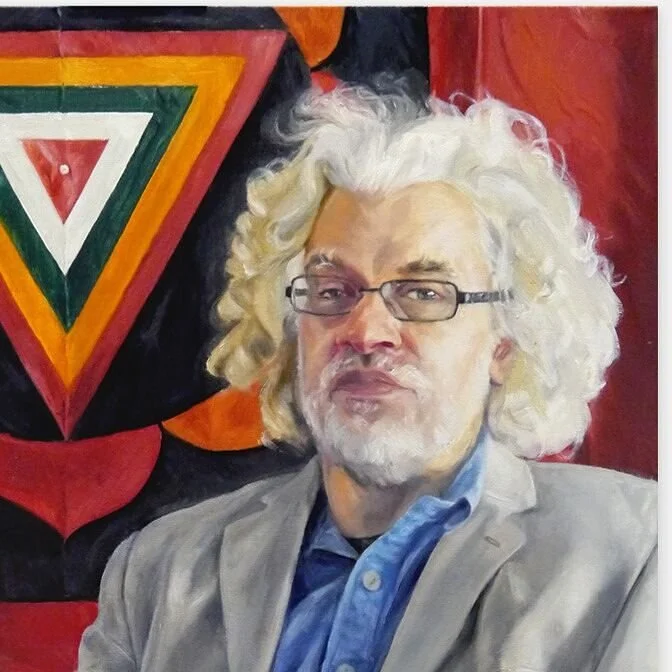 Phil Hine con cabello rizado y canoso, usando gafas, con barba, vestido con saco y camisa azul, en un fondo con un mural de colores y forma triangular.