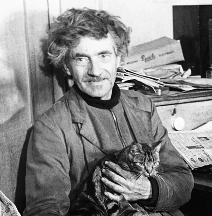 Austin Osman Spare con cabello rizado y barba sosteniendo un gato en un ambiente de interior con papeles y libros en el fondo.