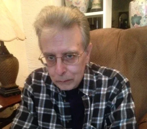 Thomas Ligotti con cabello gris, gafas, camiseta de cuadros y expresión seria, sentado en una sala con muebles y decoración en el fondo.