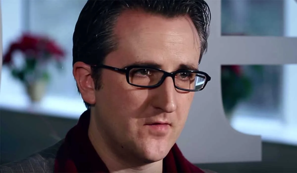 Nick Soulsby con gafas, cabello oscuro, en interior, fondo difuso con plantas y ventana.