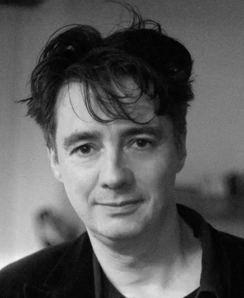 Phil Baker con cabello oscuro y ondulado, expresión calmada, en un entorno indistinto en tonos de gris.