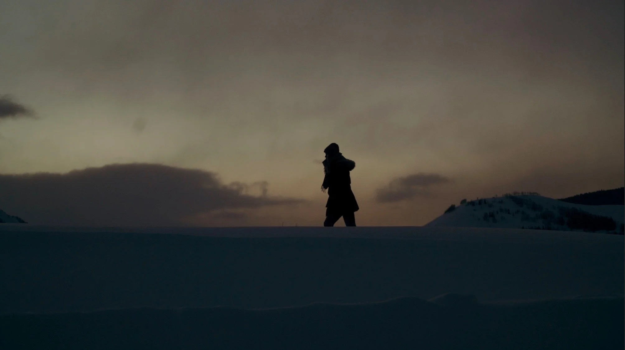 Silhouette of a person walking across snow-covered landscape during sunset or sunrise with cloudy sky in the background.