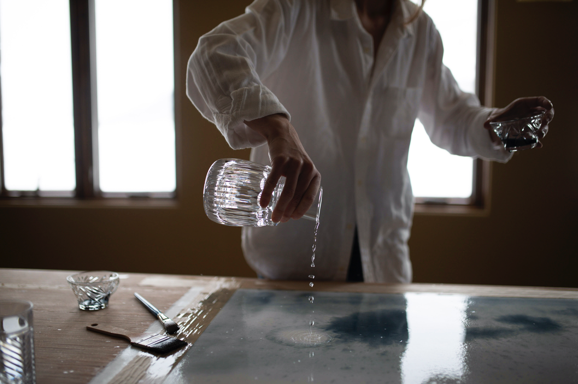 Person pouring water onto a glass tabletop while holding a glass in each hand, with paintbrush and small bowls on a wooden table in front, in front of a window.