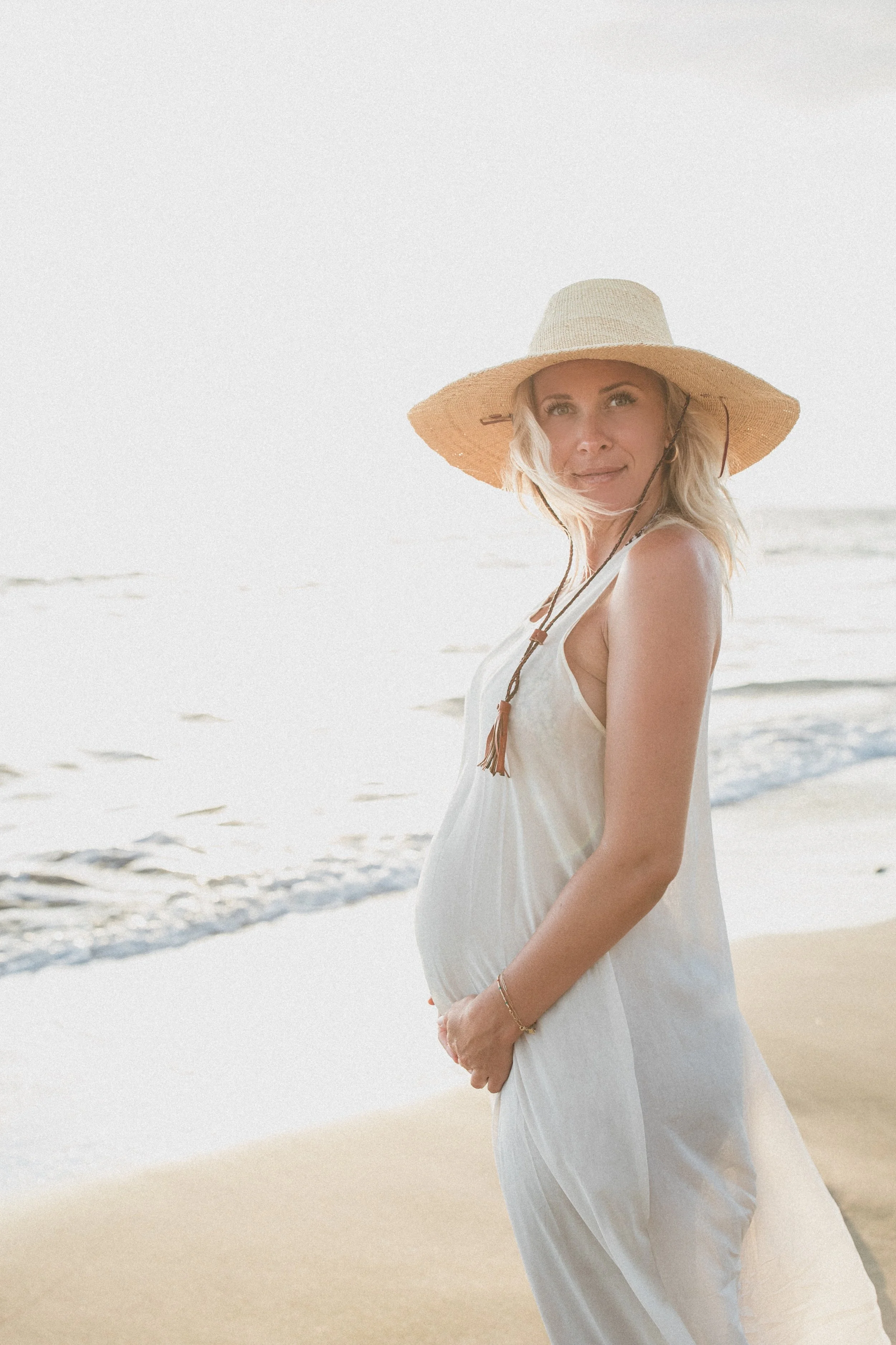 Pregnant woman in a white dress and wide-brimmed hat on the beach, looking at the camera.