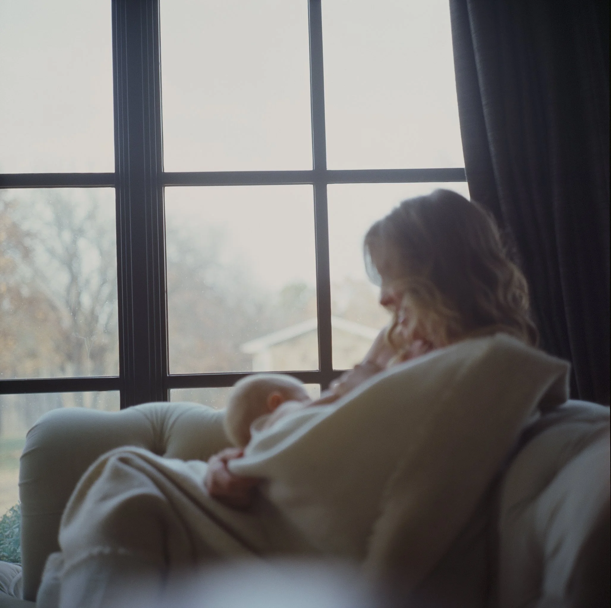 A woman with wavy hair smiling and holding a baby on a couch next to a window with curtains.