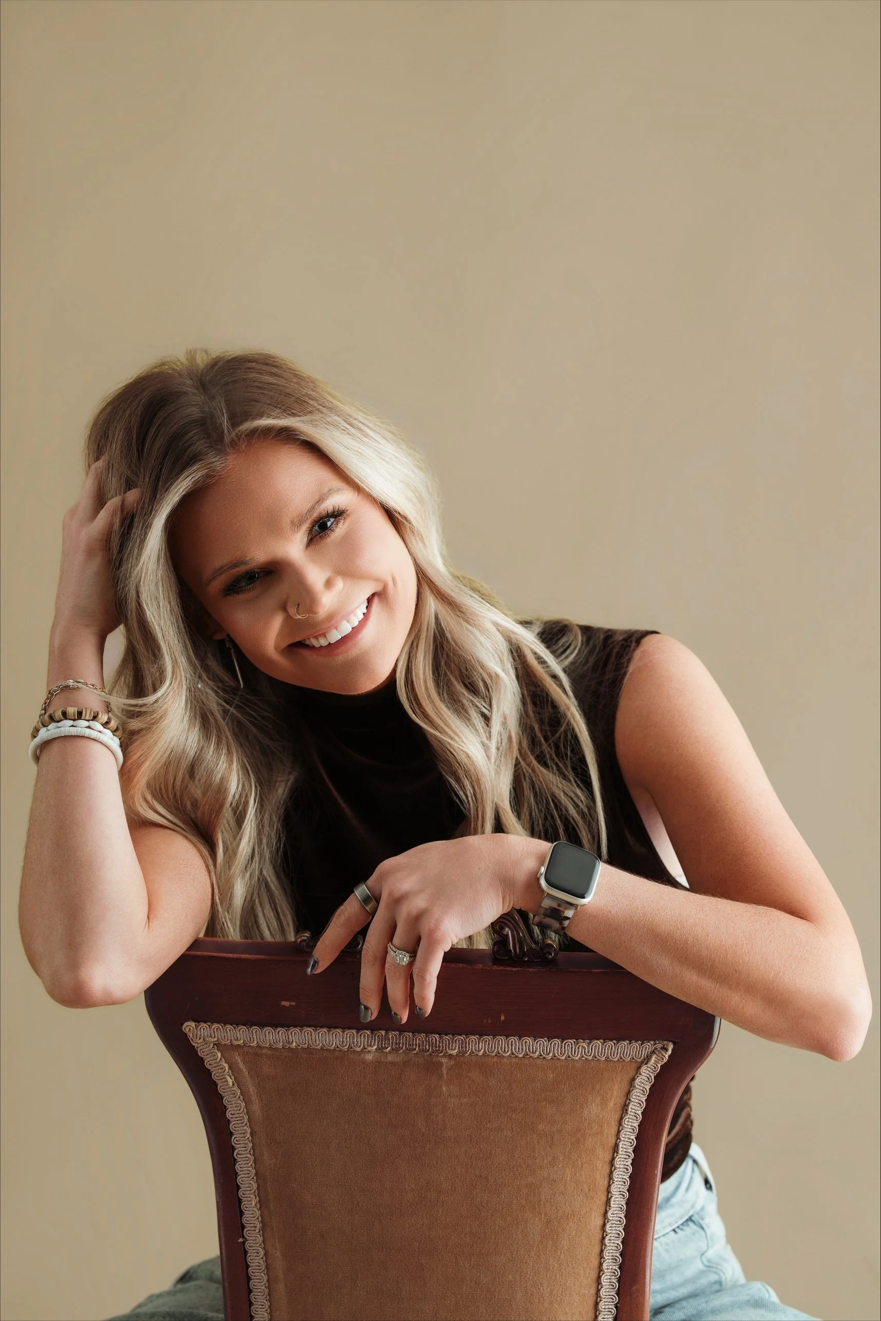 A smiling young woman with long blonde hair, wearing a black sleeveless top, jewelry, and a smartwatch, leaning on a wooden chair with a beige wall in the background.