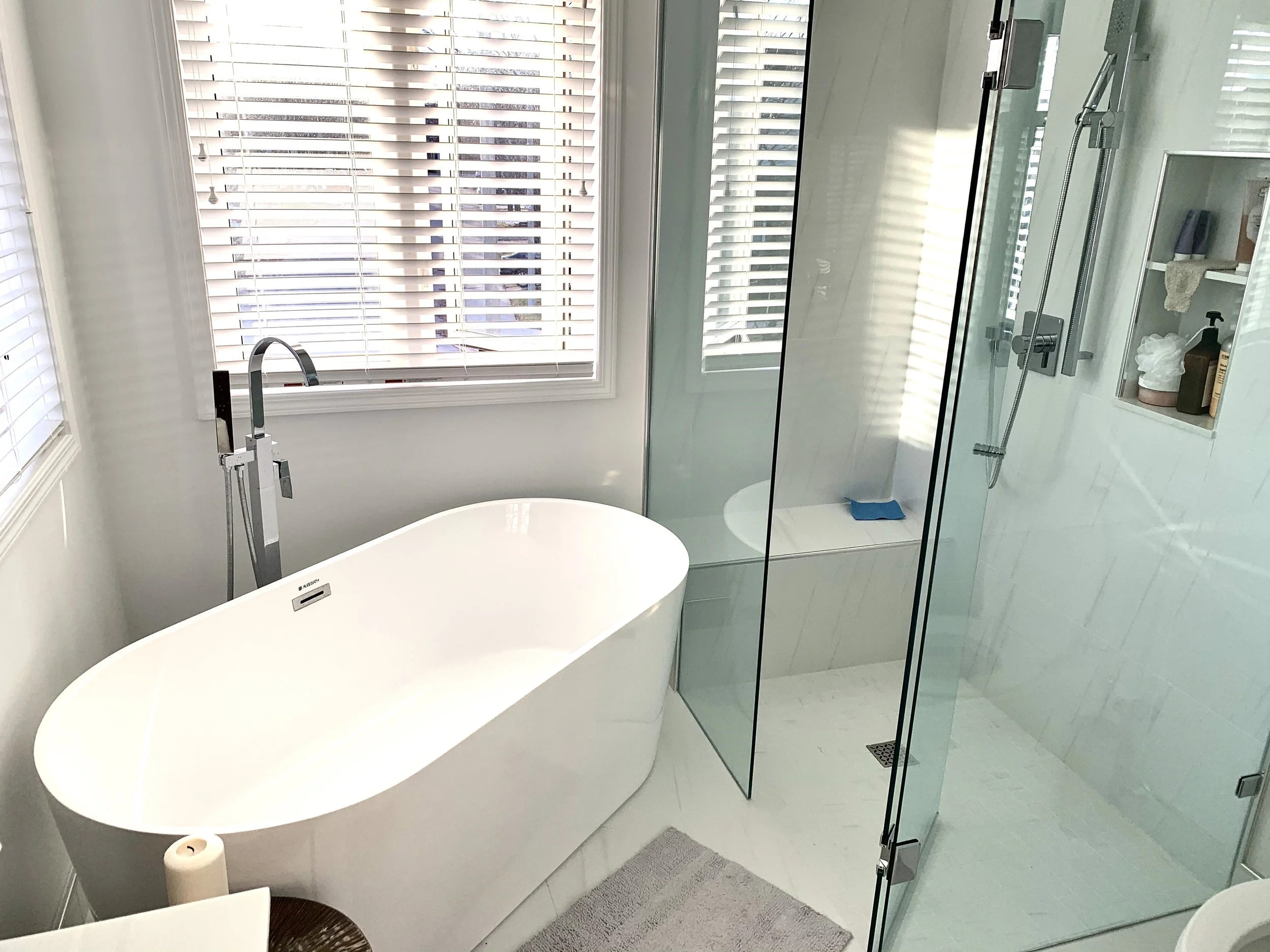 A modern bathroom with a white bathtub near a window with blinds, a glass-enclosed shower with a built-in shelf, and a gray rug on the floor.