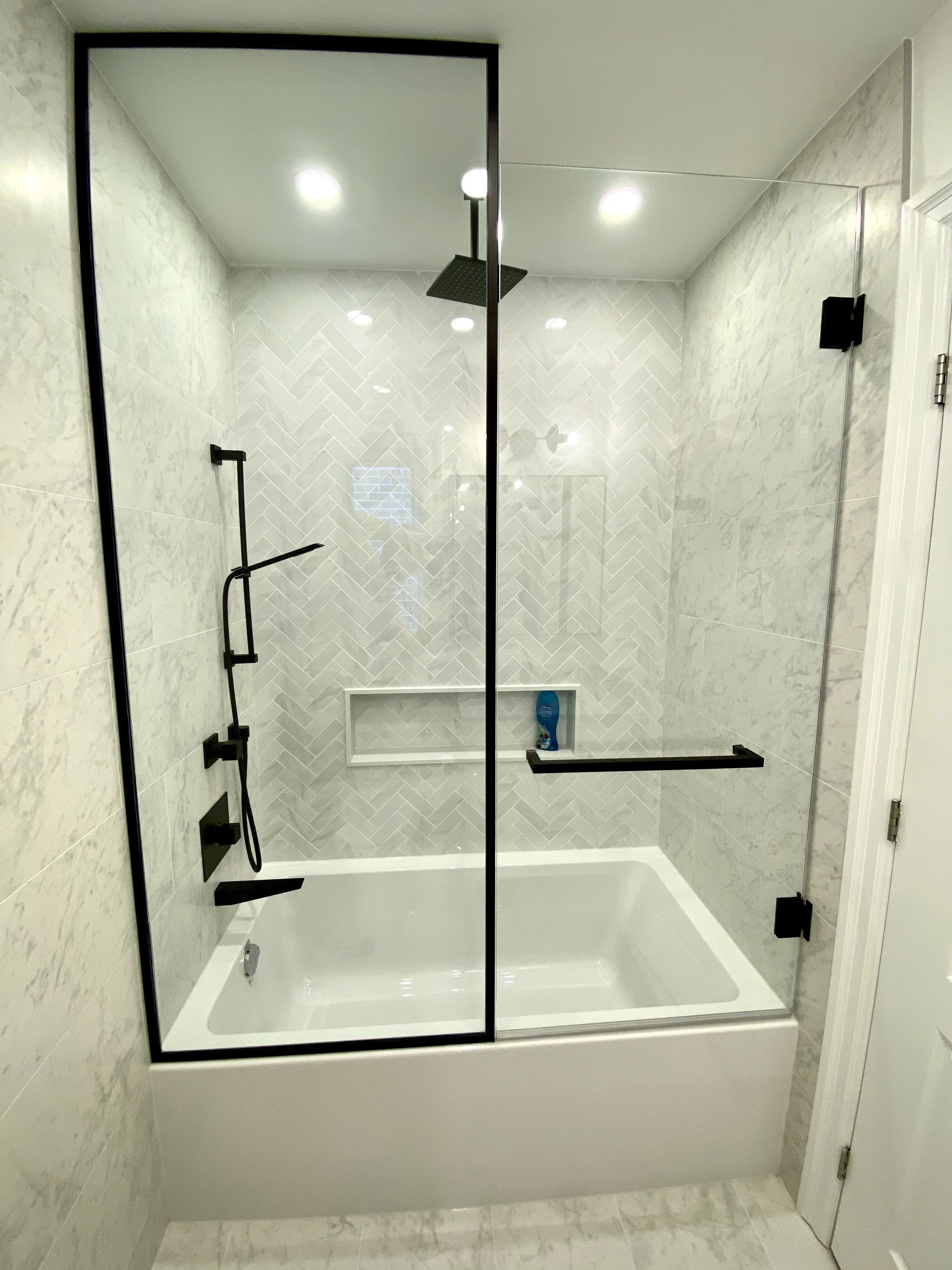 Modern bathtub and shower with black fixtures and glass enclosure in a bathroom with white marble tiles.