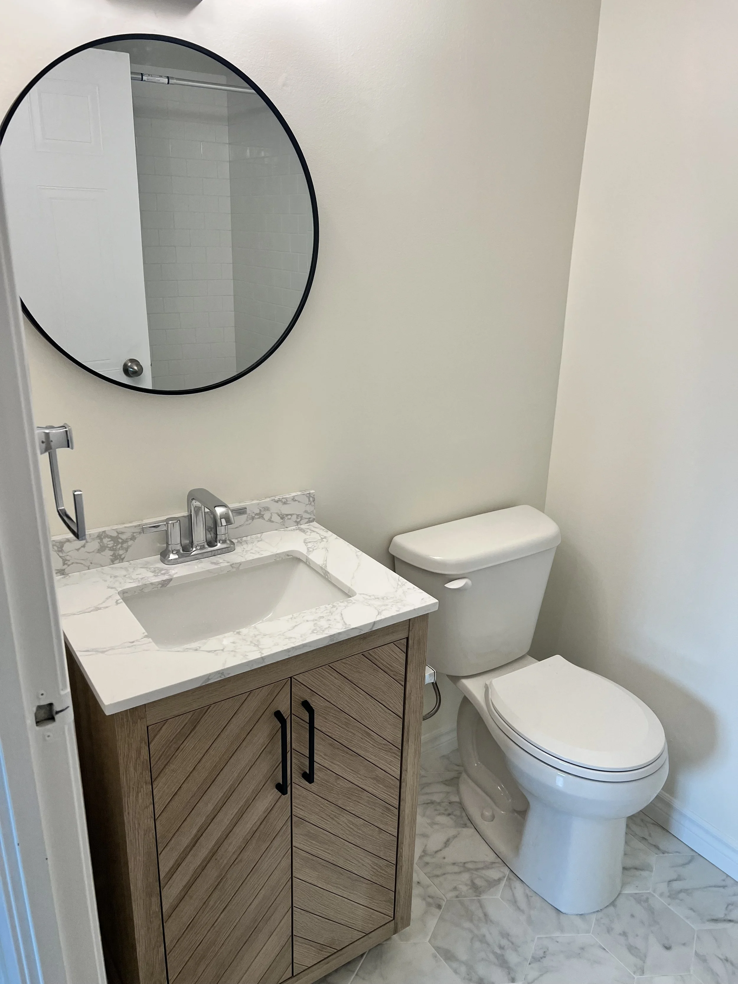 A small bathroom with a wooden cabinet and a marble countertop sink next to a toilet. A round mirror hangs above the sink, reflecting a partially open shower door with white tiled walls. The floor is tiled in a white marble pattern.