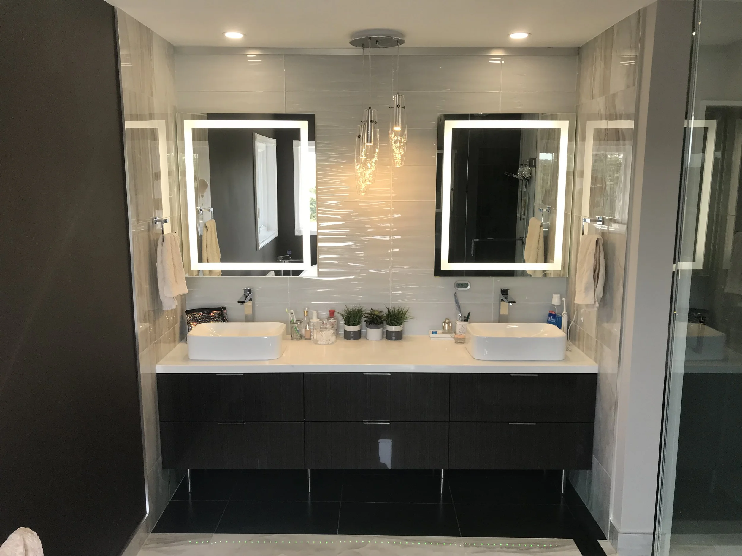 Modern bathroom with double vanity featuring two rectangular vessel sinks, illuminated mirrors, and three small potted plants on the countertop. Towel holders with towels, and bathroom toiletries are also visible.