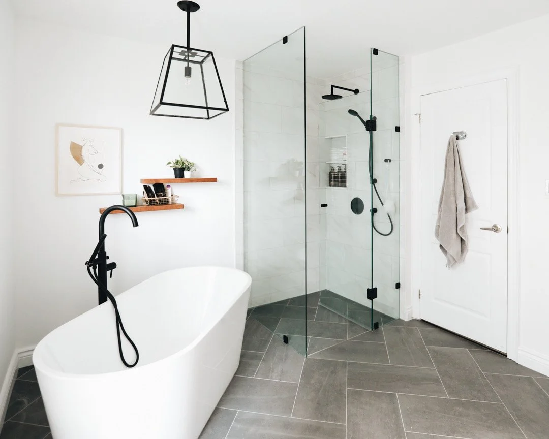 Modern bathroom with a white freestanding bathtub, black wall-mounted faucet, and a walk-in glass shower with black fixtures, grey tile flooring, and a white wall with two wooden shelves holding decor and toiletries.