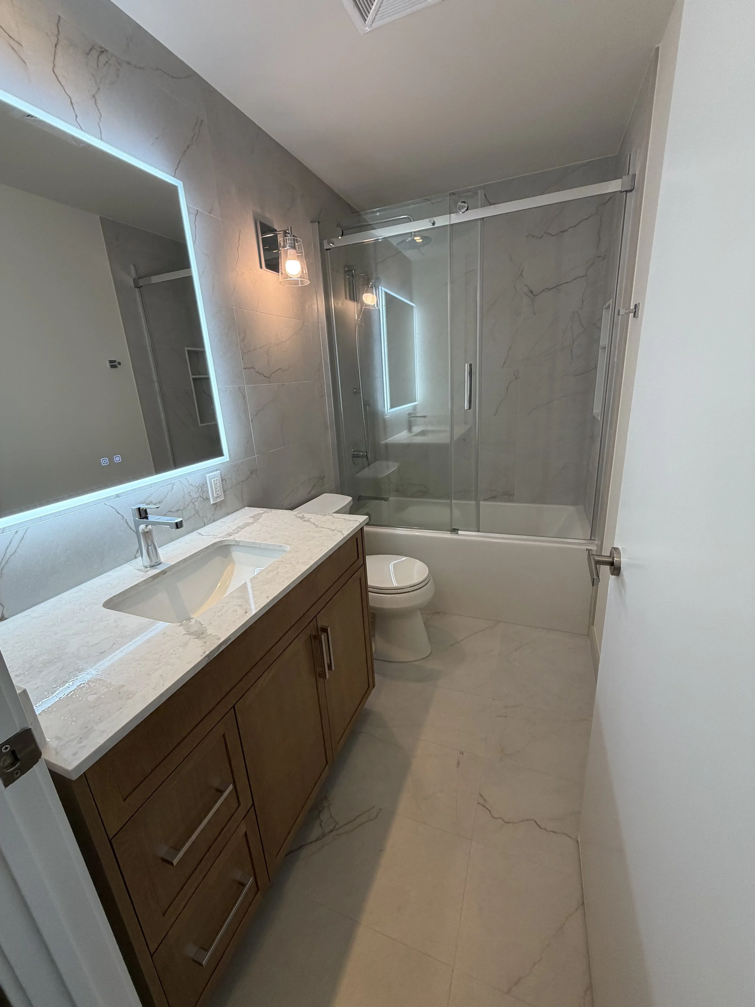 Modern bathroom with a marble vanity top, wooden cabinet, wall-mounted mirror with LED lighting, glass shower with marble walls, and a white toilet.