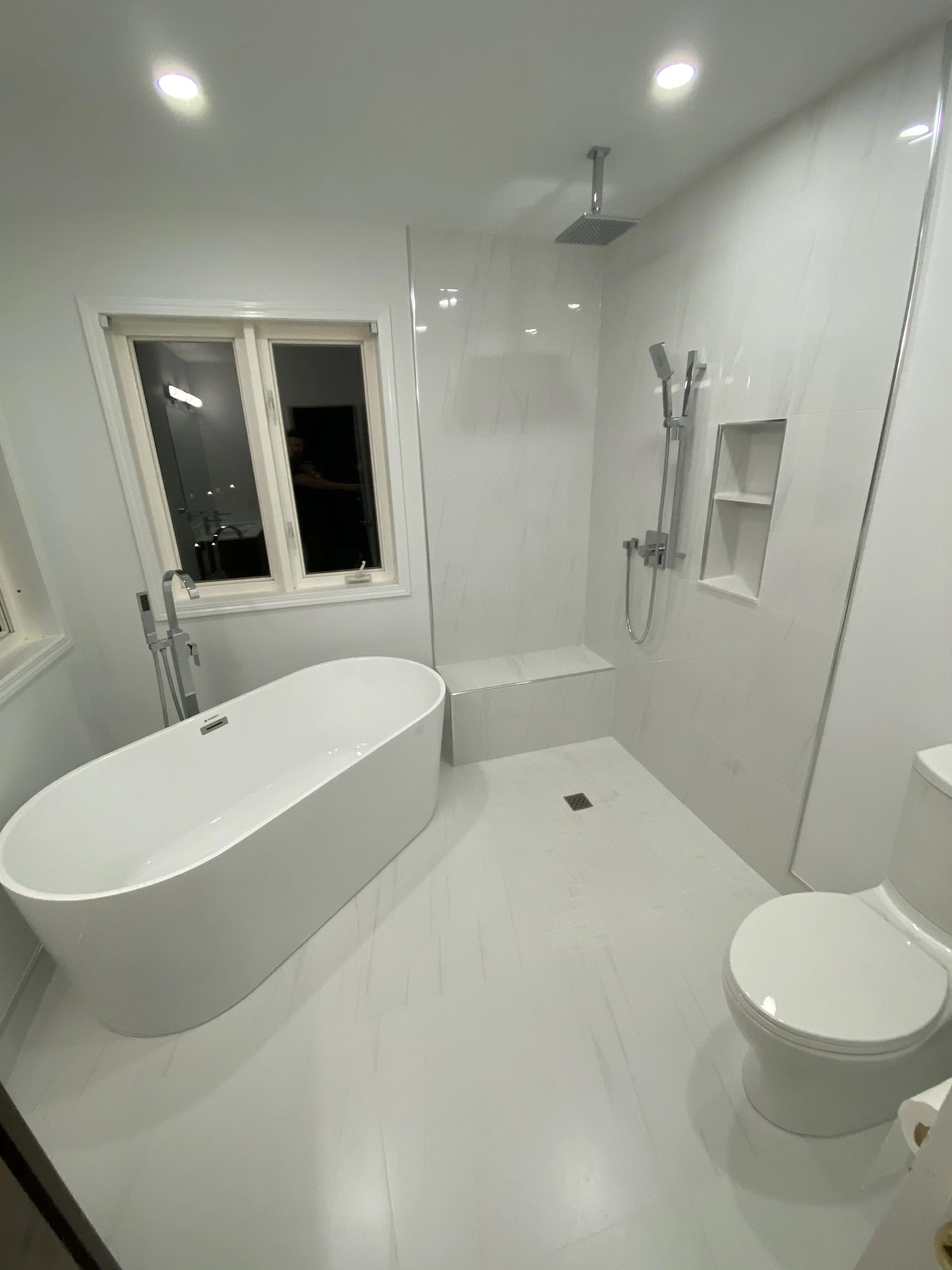 Modern bathroom with white walls, a white bathtub, a window, a walk-in shower, and a toilet.