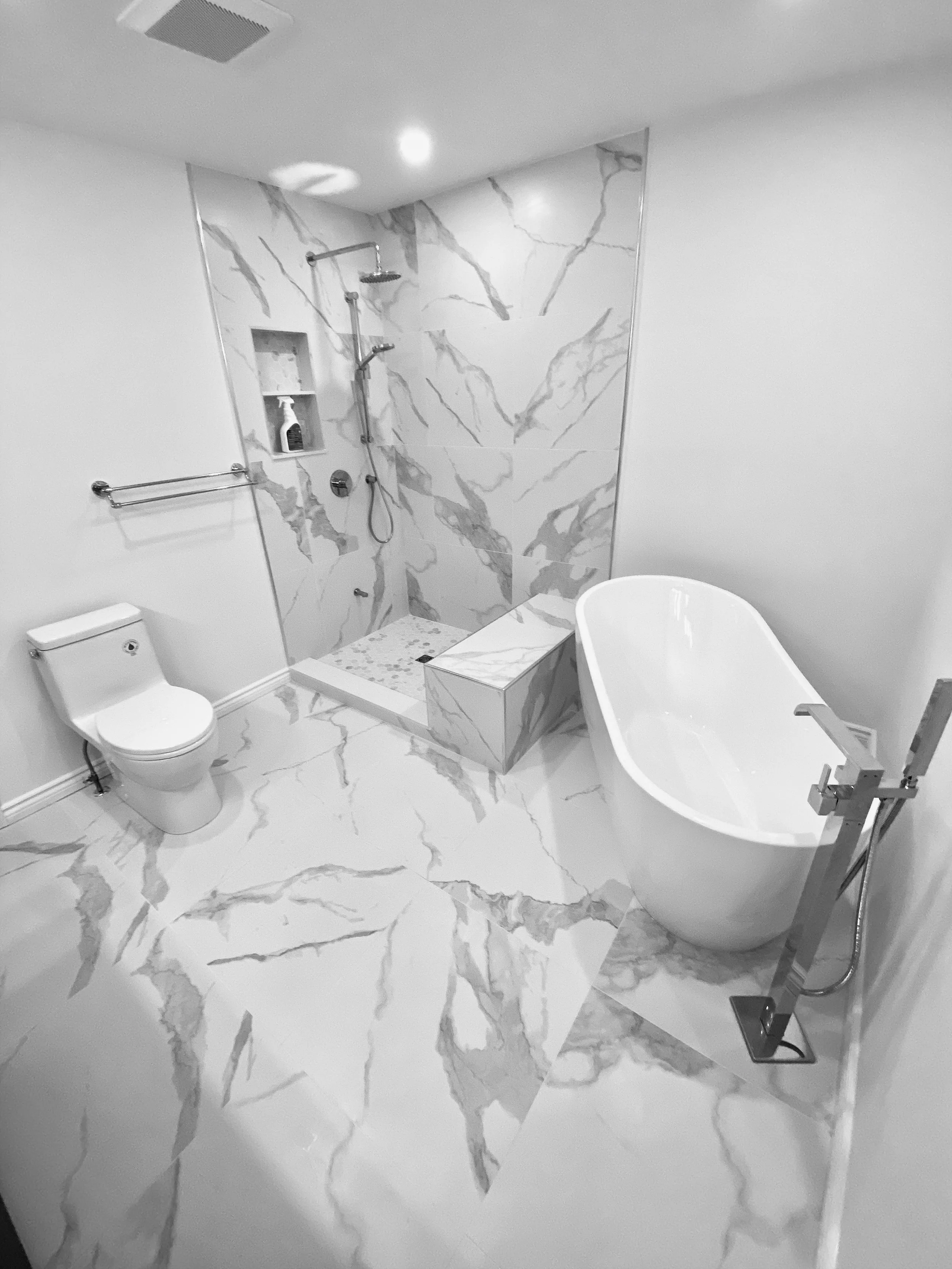 A modern bathroom with marble flooring and marble accent wall in the shower area. It features a toilet on the left, a minimalist bathtub on the right, and a walk-in shower with a built-in shelf in the back wall.