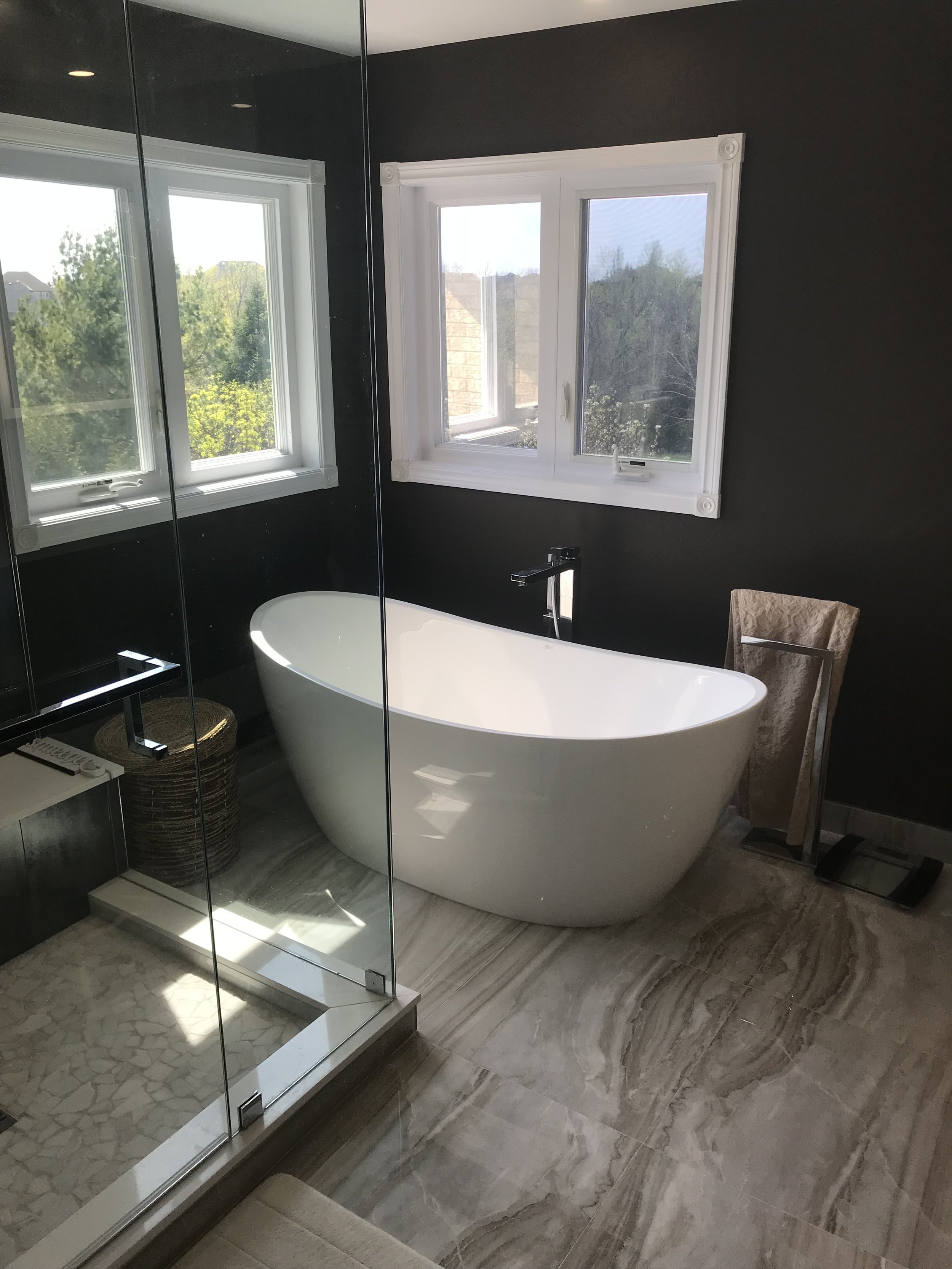 Modern bathroom with black wall, white freestanding bathtub, and large windows showing trees outside.