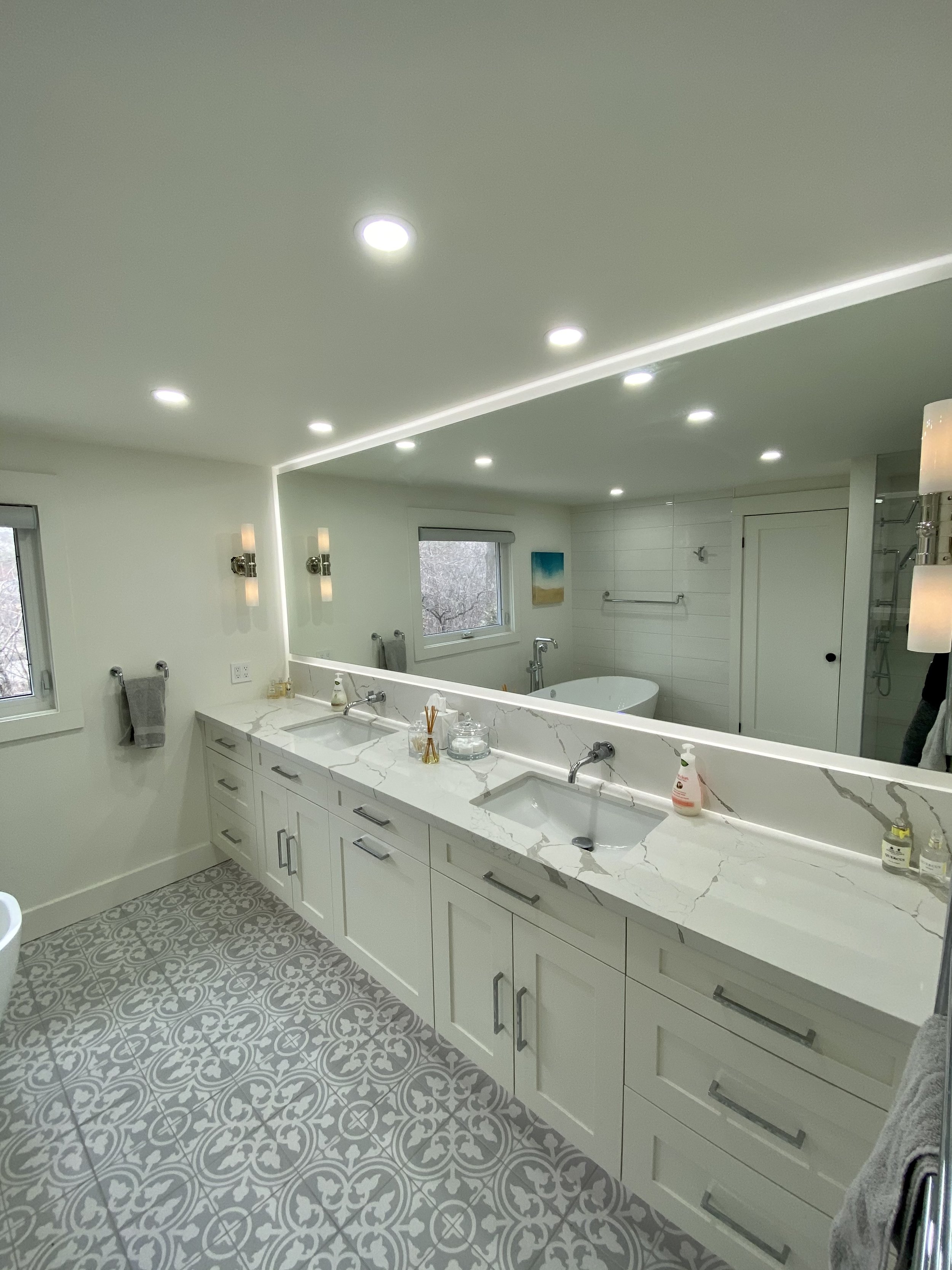 Modern bathroom with white vanity, marble countertop, and decorative tile floor, featuring a large mirror, double sinks, and a view into a shower area.