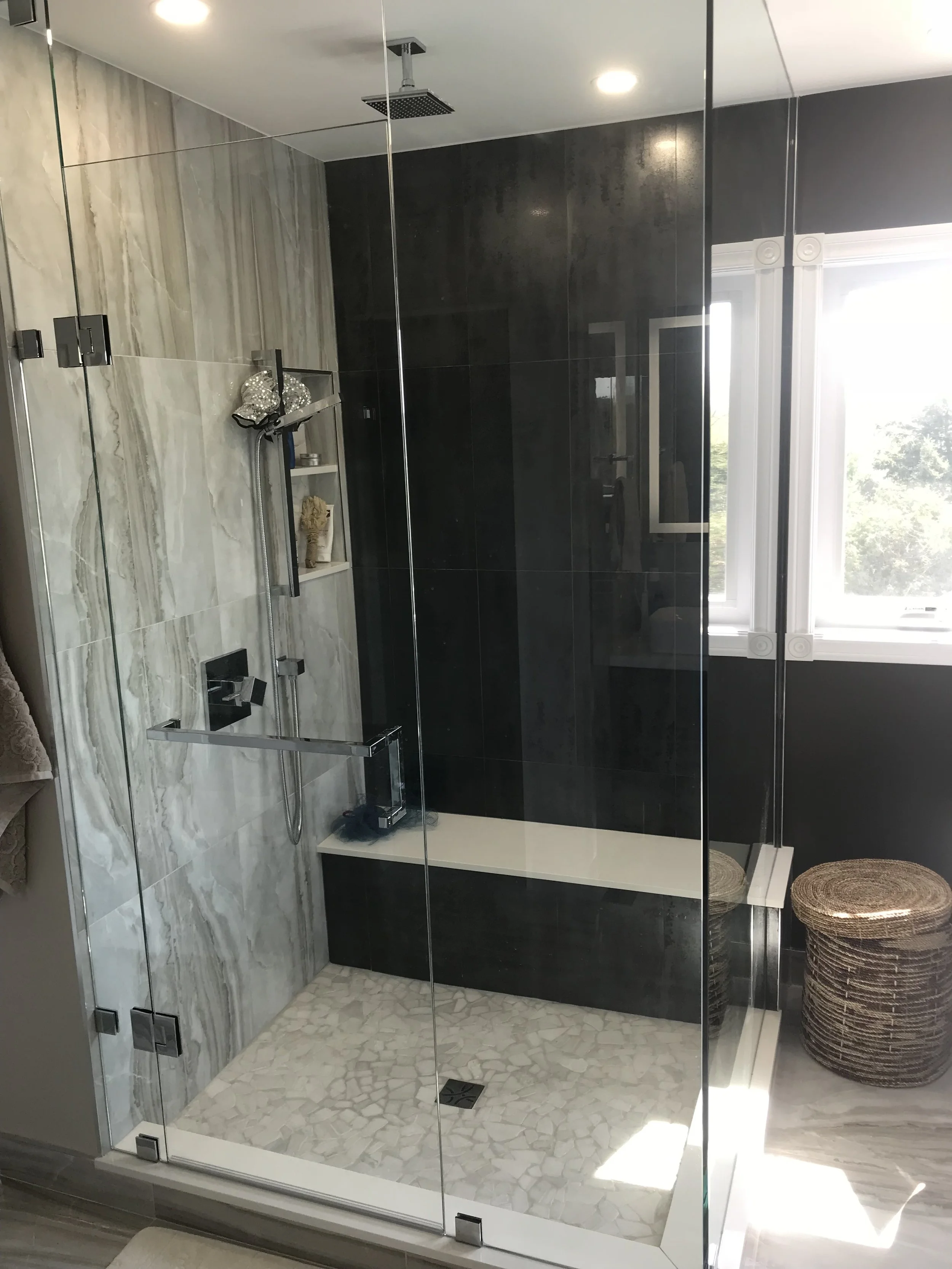 A modern glass-enclosed shower with beige and black tile walls and a pebble floor, mounted shower fixtures, and a window with a white frame.