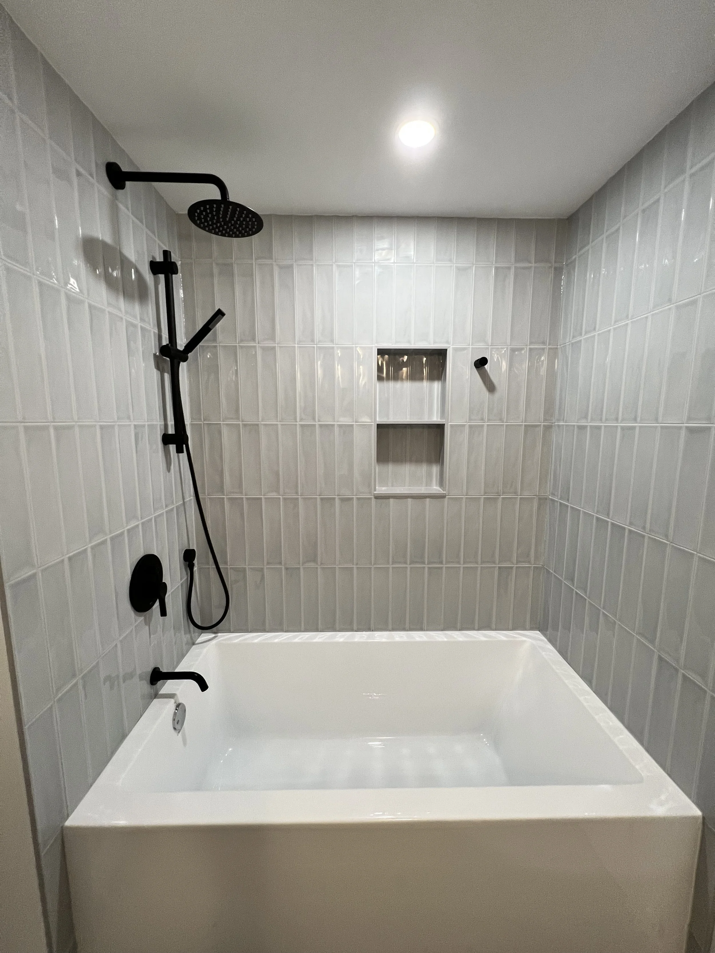 Bathroom with white tiled walls, black shower fixtures, and a white bathtub under a ceiling light.