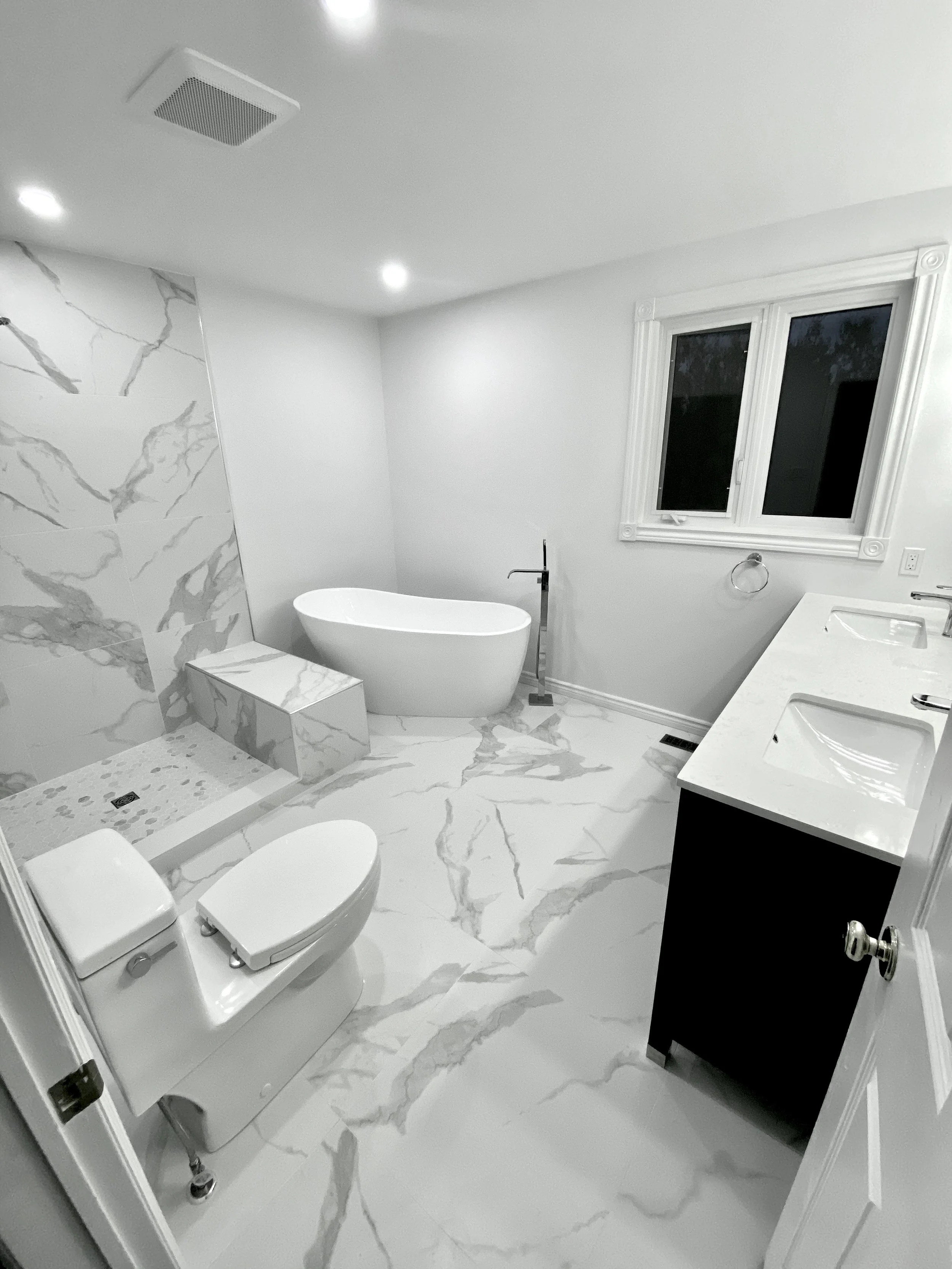 Modern bathroom with a white bathtub, a toilet, a black and white vanity with two sinks, a window, and marble floors and wall accent.