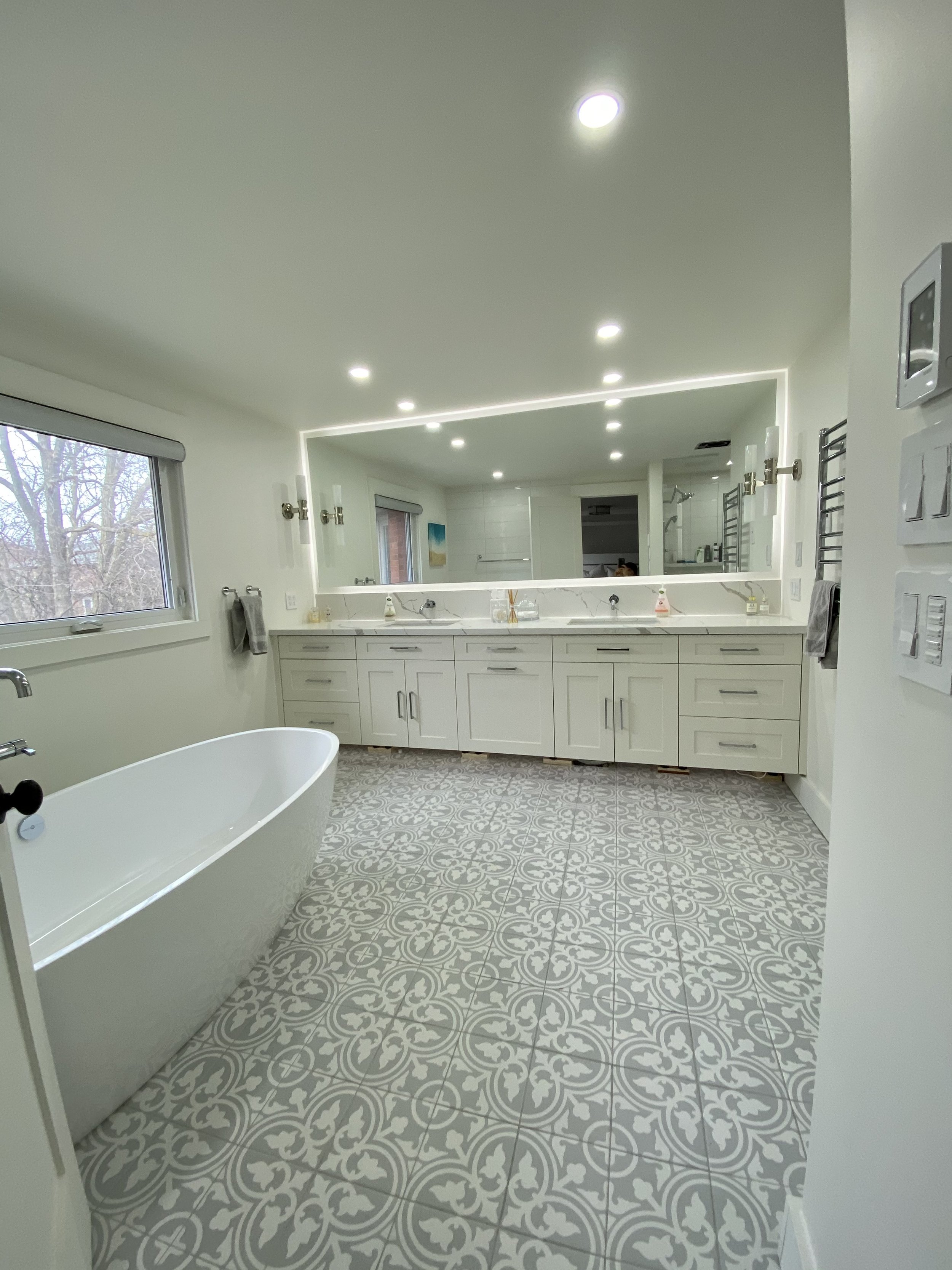 Modern bathroom with white cabinetry, large mirror, bathtub, patterned floor tiles, and two windows.