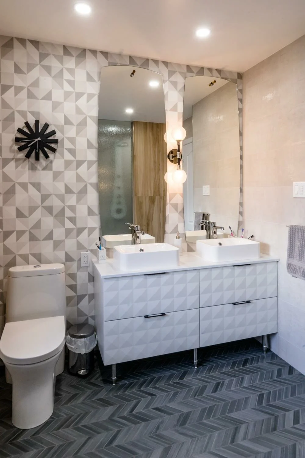 Modern bathroom with geometric patterned wall tiles, a large mirror, two white vessel sinks on a vanity, wall-mounted light fixture, toilet, and dark herringbone floor tiles.