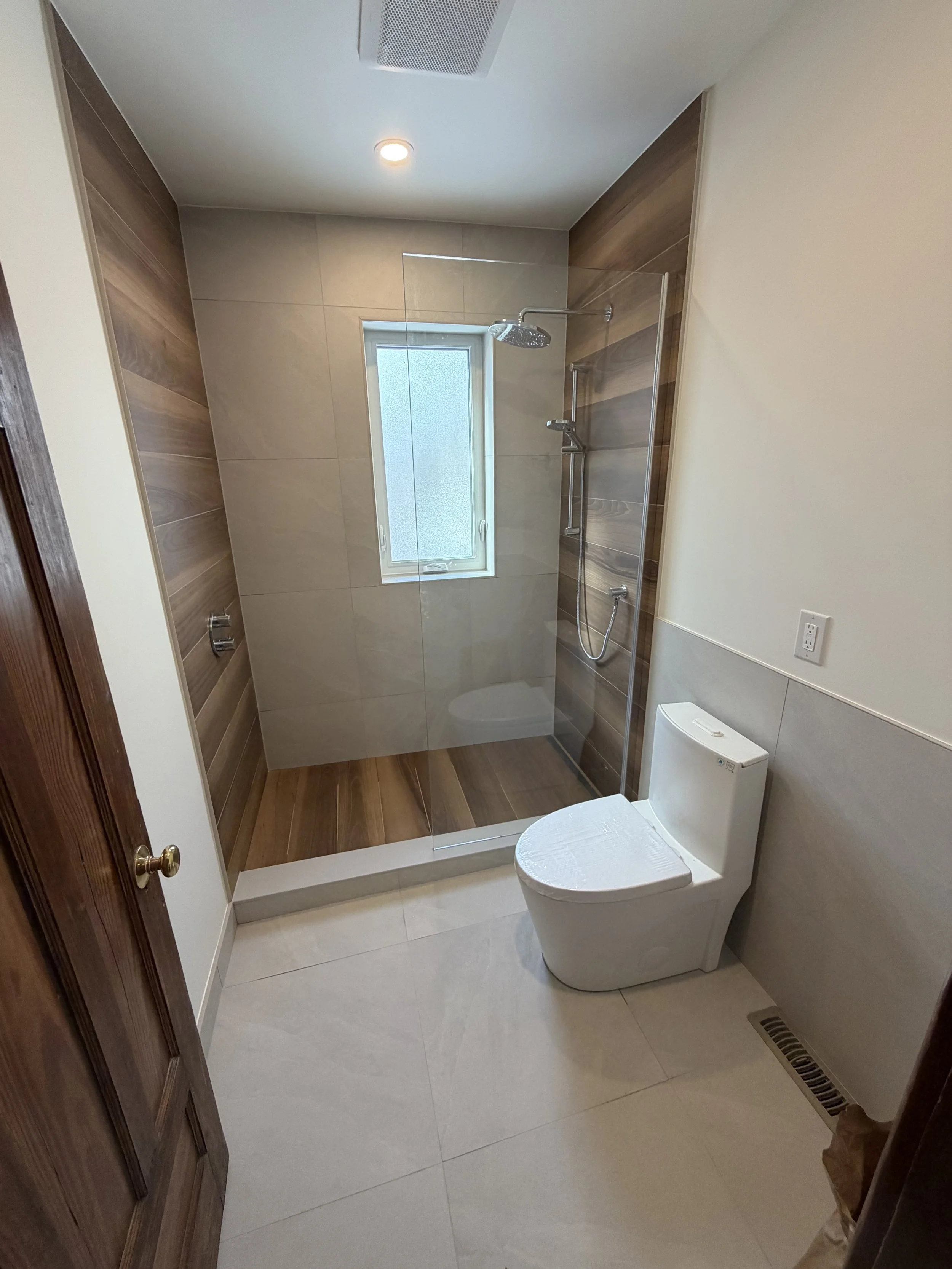 Bathroom with a walk-in shower featuring a glass door, a large window, and wood-like wall and floor tiles. Toilet in the foreground and a wooden door on the side.