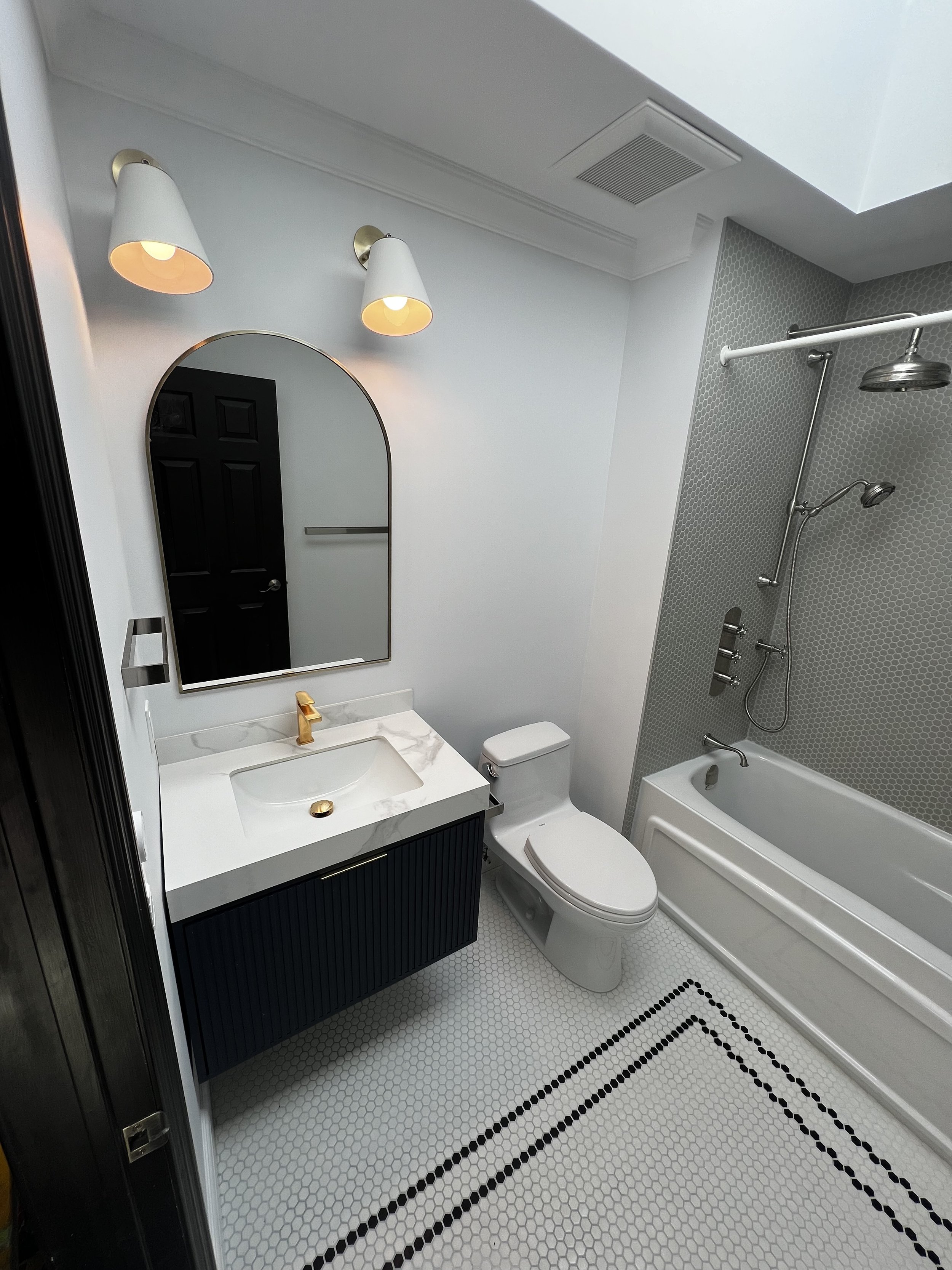 Bathroom with white and black vanity, gold faucet, oval mirror, wall-mounted sconces, white toilet, bathtub with shower head, hexagon tile floor.