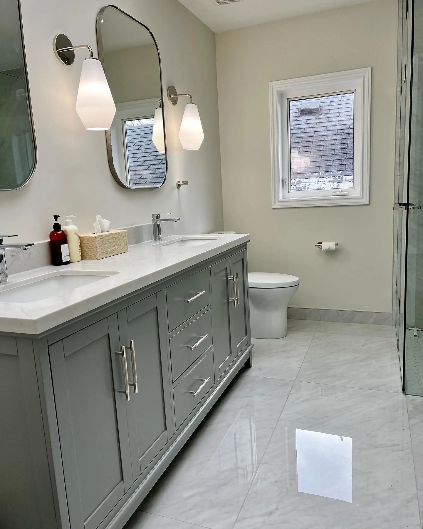 A modern bathroom with a double vanity with a marble countertop, two mirrors, wall-mounted lights, a toilet, a window, and a glass-enclosed shower.