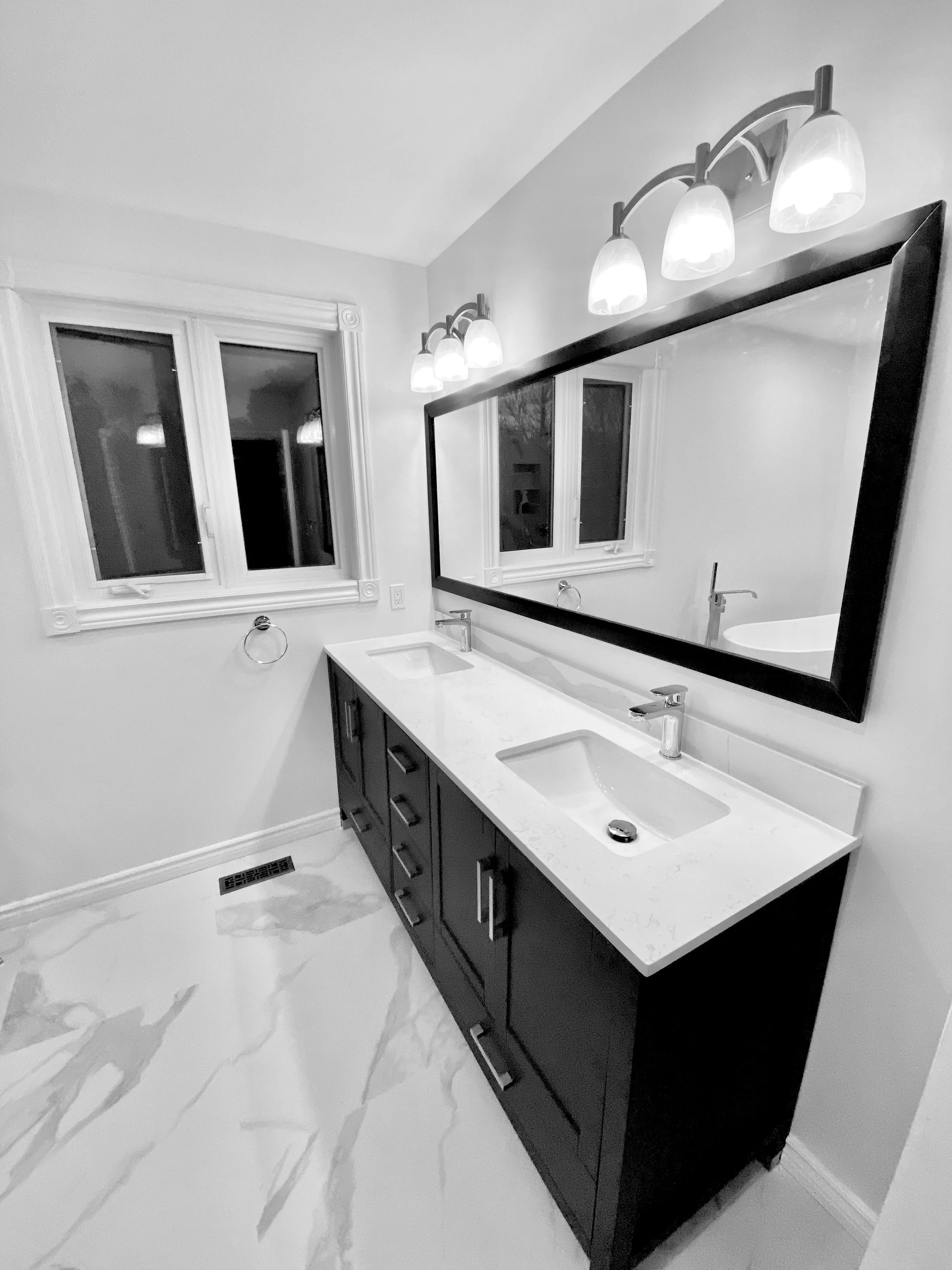 A modern bathroom with a double sink vanity, large mirror, and three-light fixture above it. The bathroom has white walls, white marble flooring, and a window with white trim.