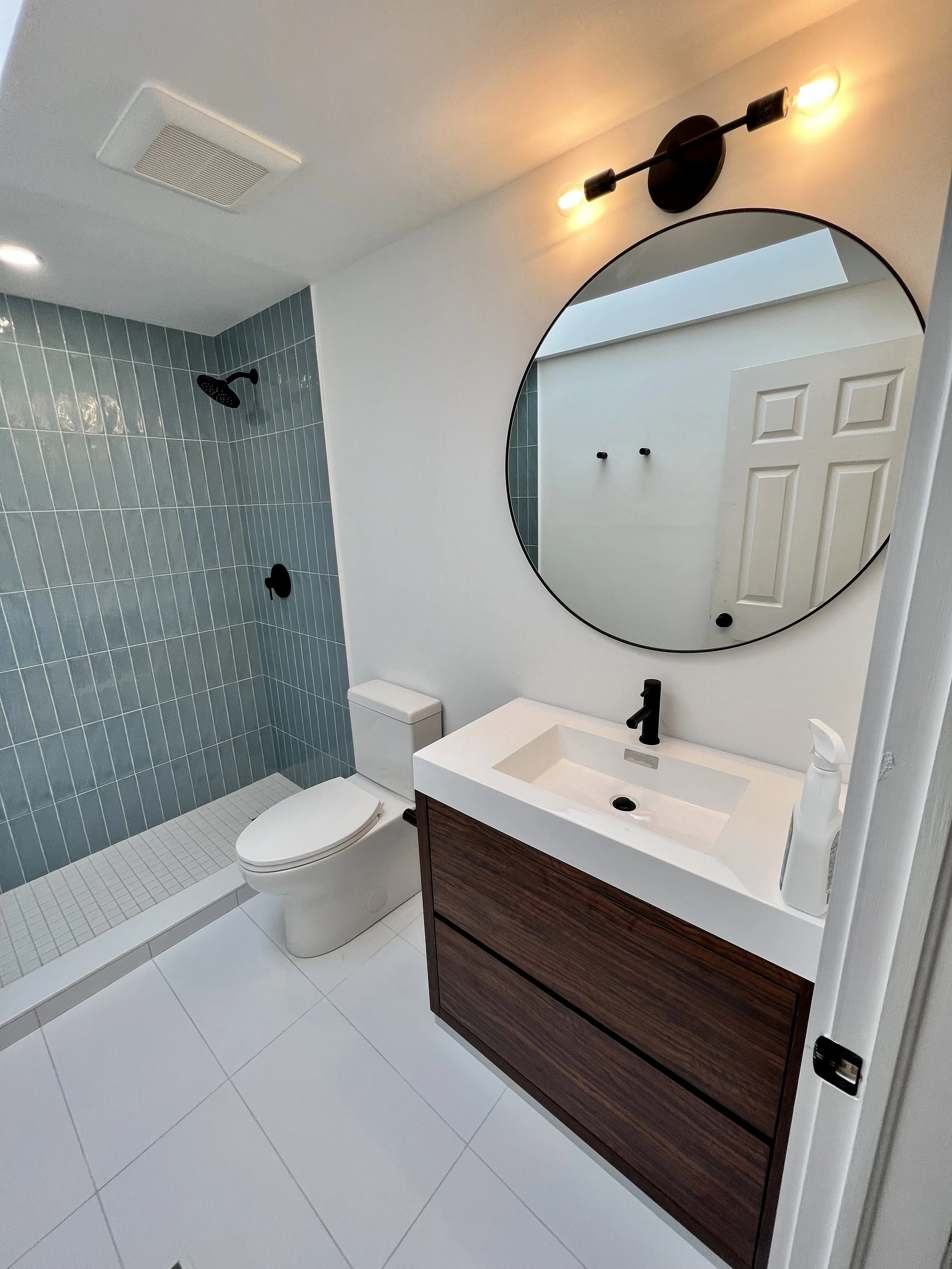 Modern bathroom with blue tiled shower, white toilet, and a wooden vanity with a white sink, round mirror, and black fixtures.