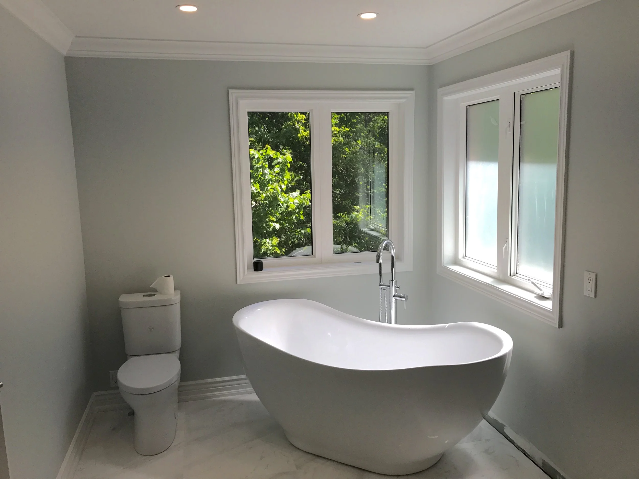 Modern bathroom with a white freestanding bathtub, a white toilet, and two large windows with green foliage outside.