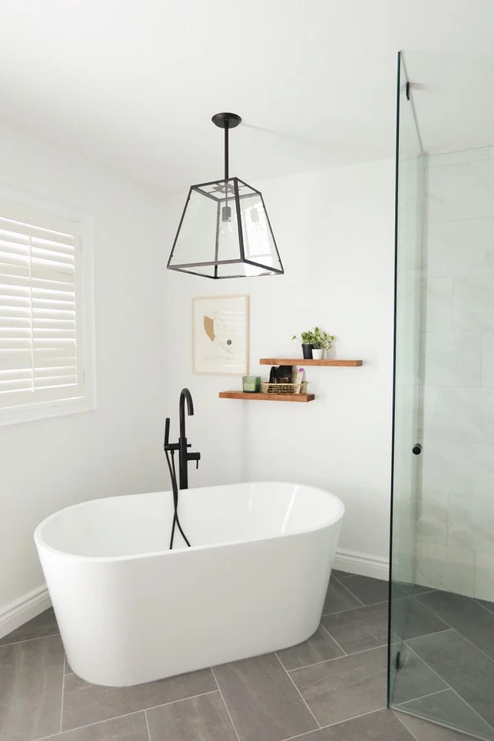 Modern bathroom with a free-standing bathtub, black faucet, white walls, a window with shutters, two wooden shelves with plants and decor, framed artwork, and a glass shower door.