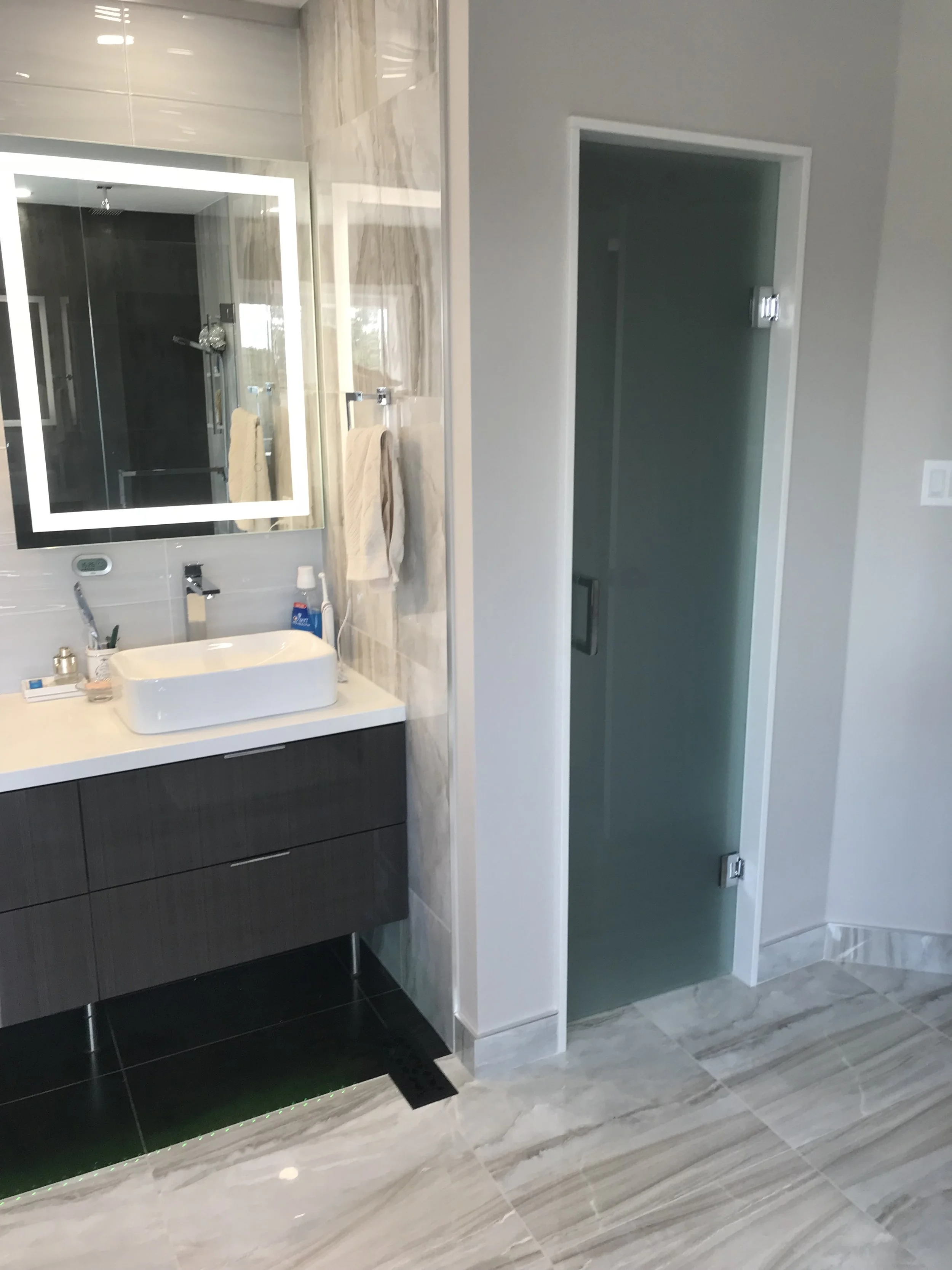 Modern bathroom with a vanity mirror, sink, towels, cleaning supplies, and a frosted glass shower enclosure.