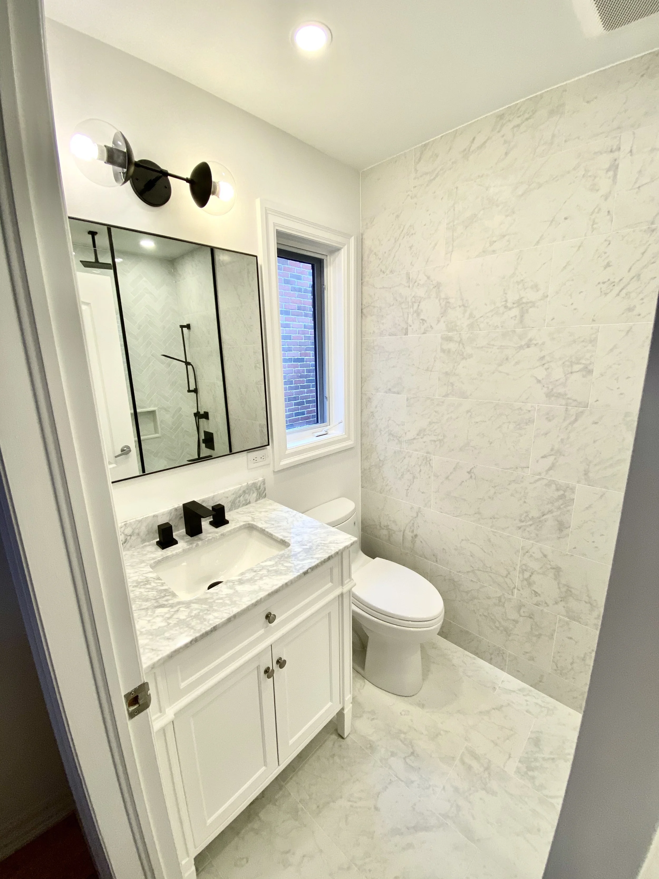 Modern bathroom with white marble vanity, black fixtures, large mirror, white toilet, window, and marble tile walls and floor.
