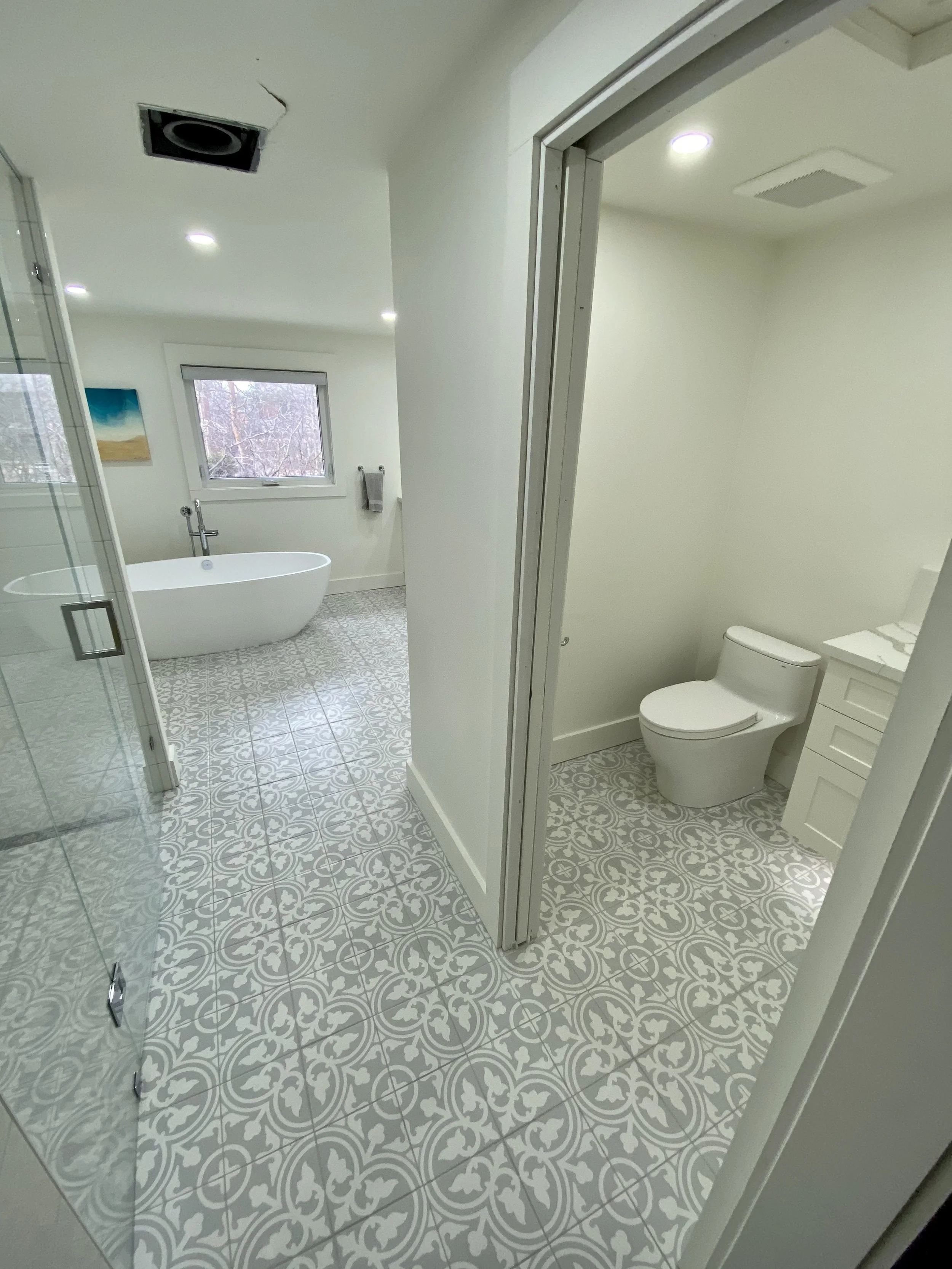 A modern bathroom with a freestanding bathtub near a window, a separate toilet room, and patterned floor tiles.