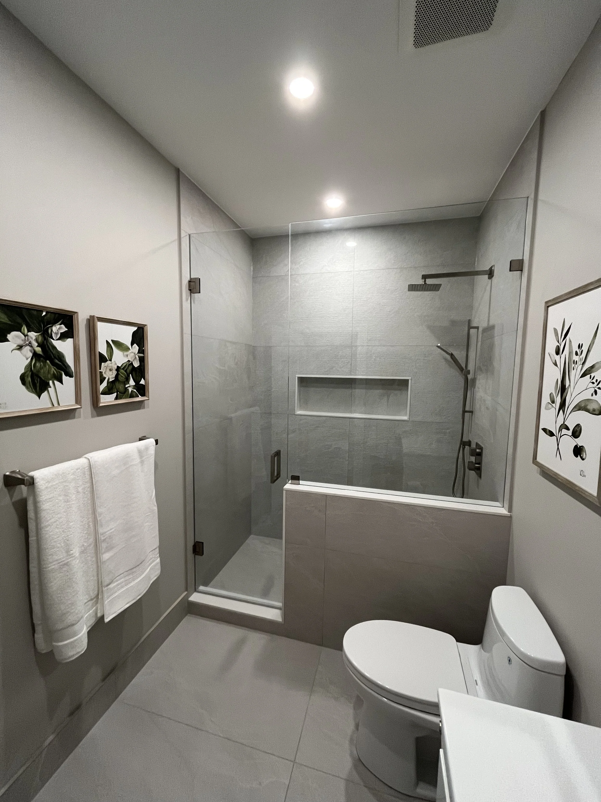 Modern bathroom with a walk-in shower, gray tiled walls, a white toilet, and framed botanical art on the walls.