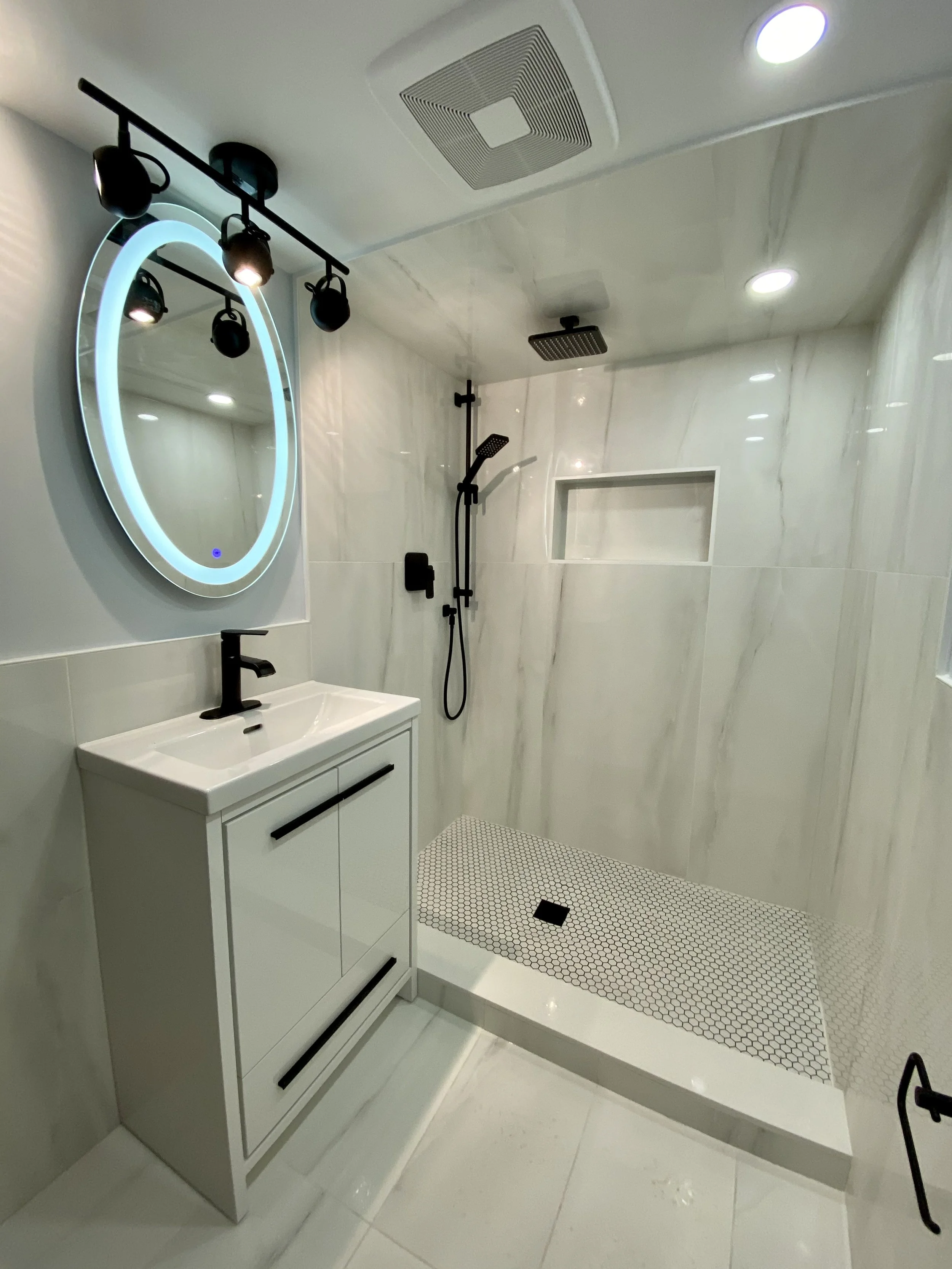 Modern bathroom with a vanity, circular LED mirror, black fixtures, and a walk-in shower with black fixtures, a built-in shelf, and small hexagon floor tiles.